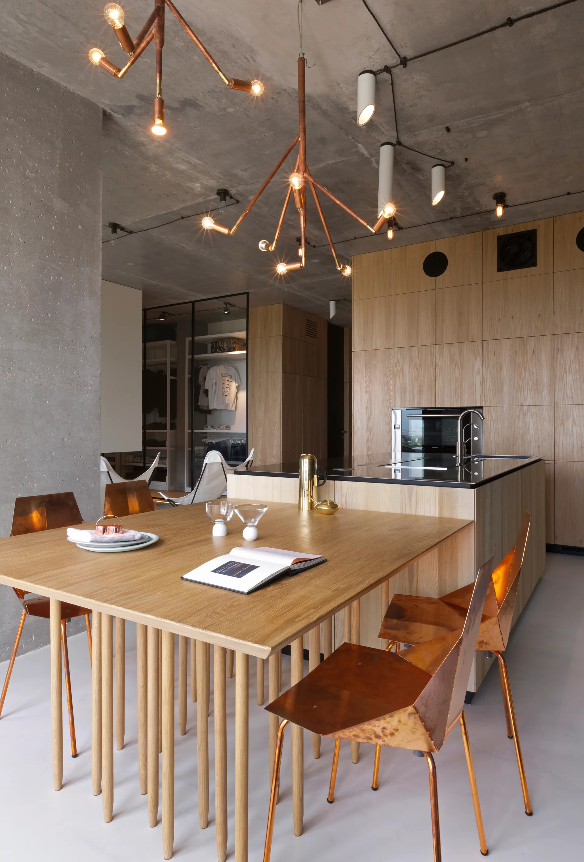 An industrial dining area with a wood table, geometric copper chairs, and a branch-like copper chandelier beneath a raw concrete ceiling.
