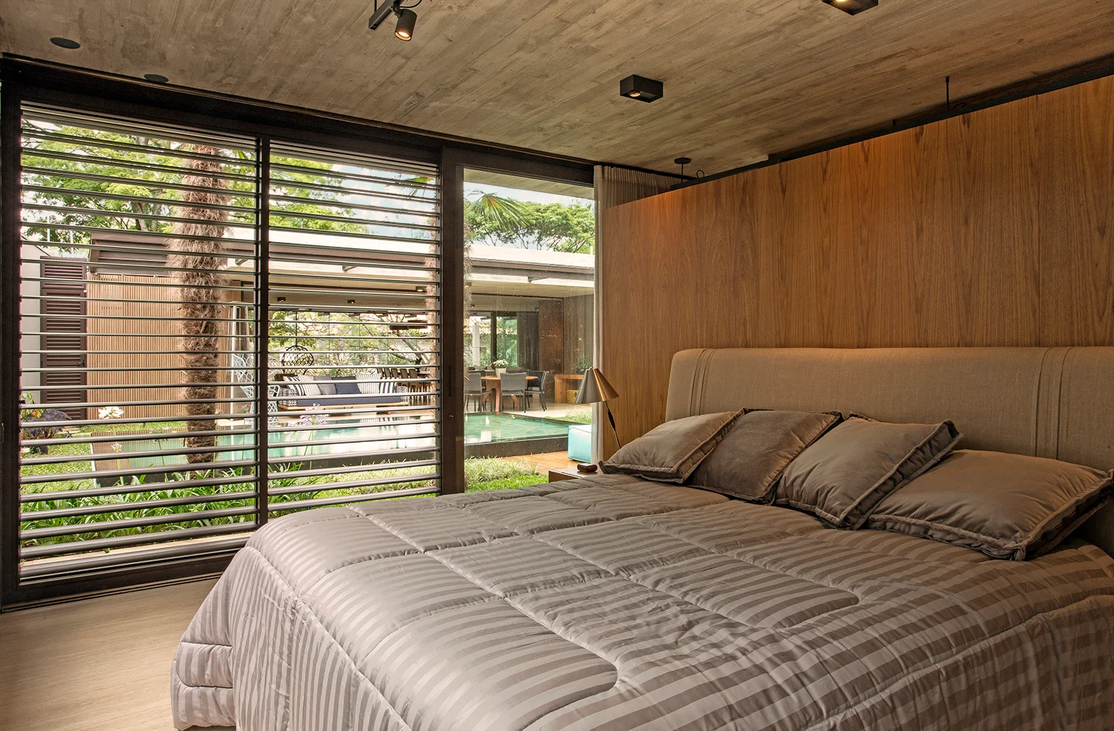 A contemporary bedroom features a large bed with gray bedding, a wood-paneled partition wall, and an exposed concrete ceiling.