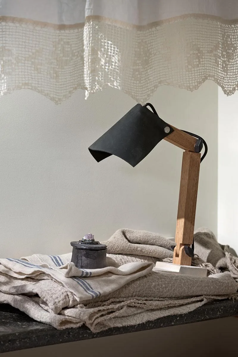 An articulating wooden desk lamp with a black shade sits on folded beige fabrics next to a small dark container beneath a white lace curtain.