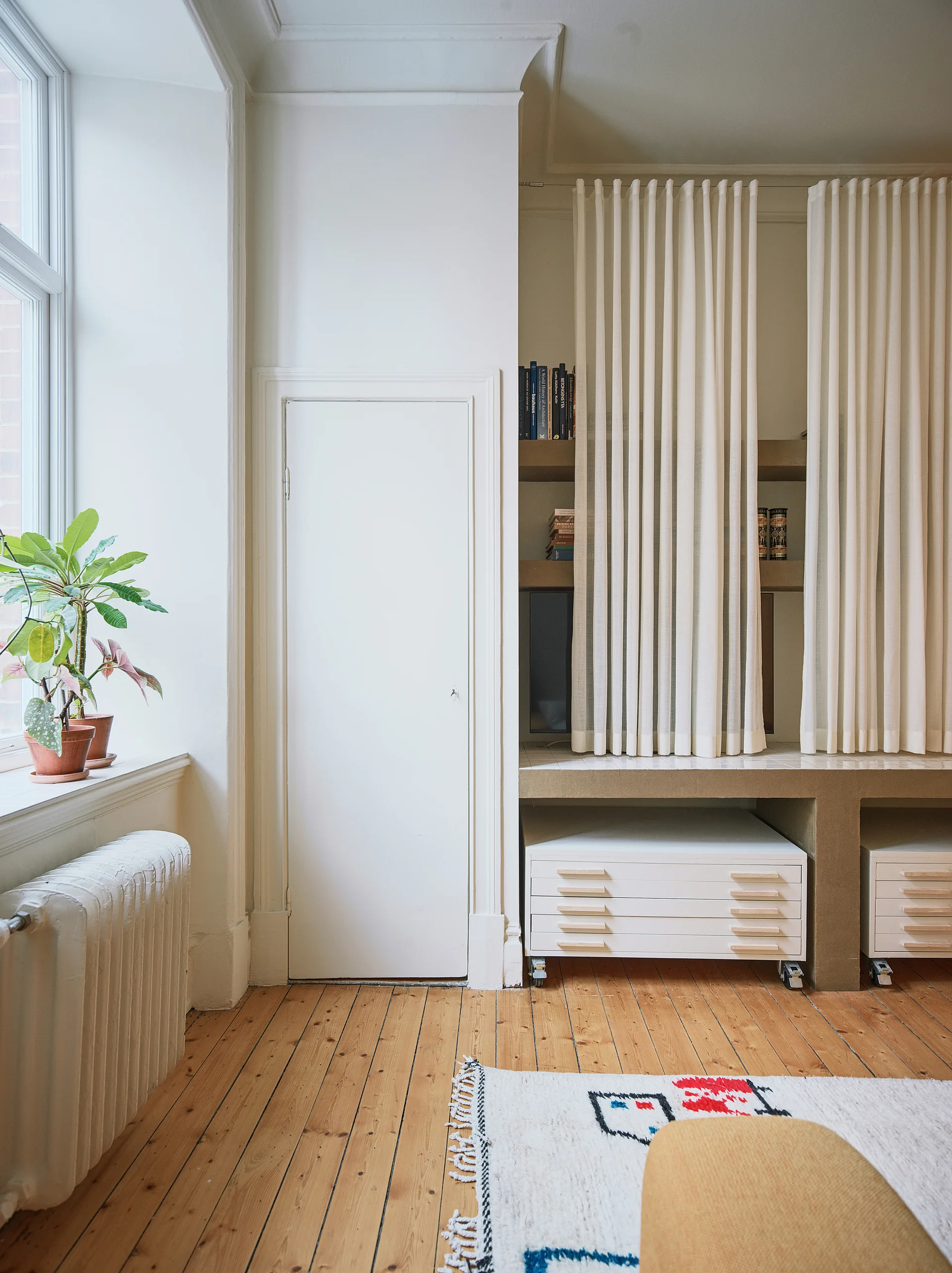A minimalist room featuring built-in shelving concealed by white fabric curtains, white rolling flat drawer cabinets, and natural wood flooring.