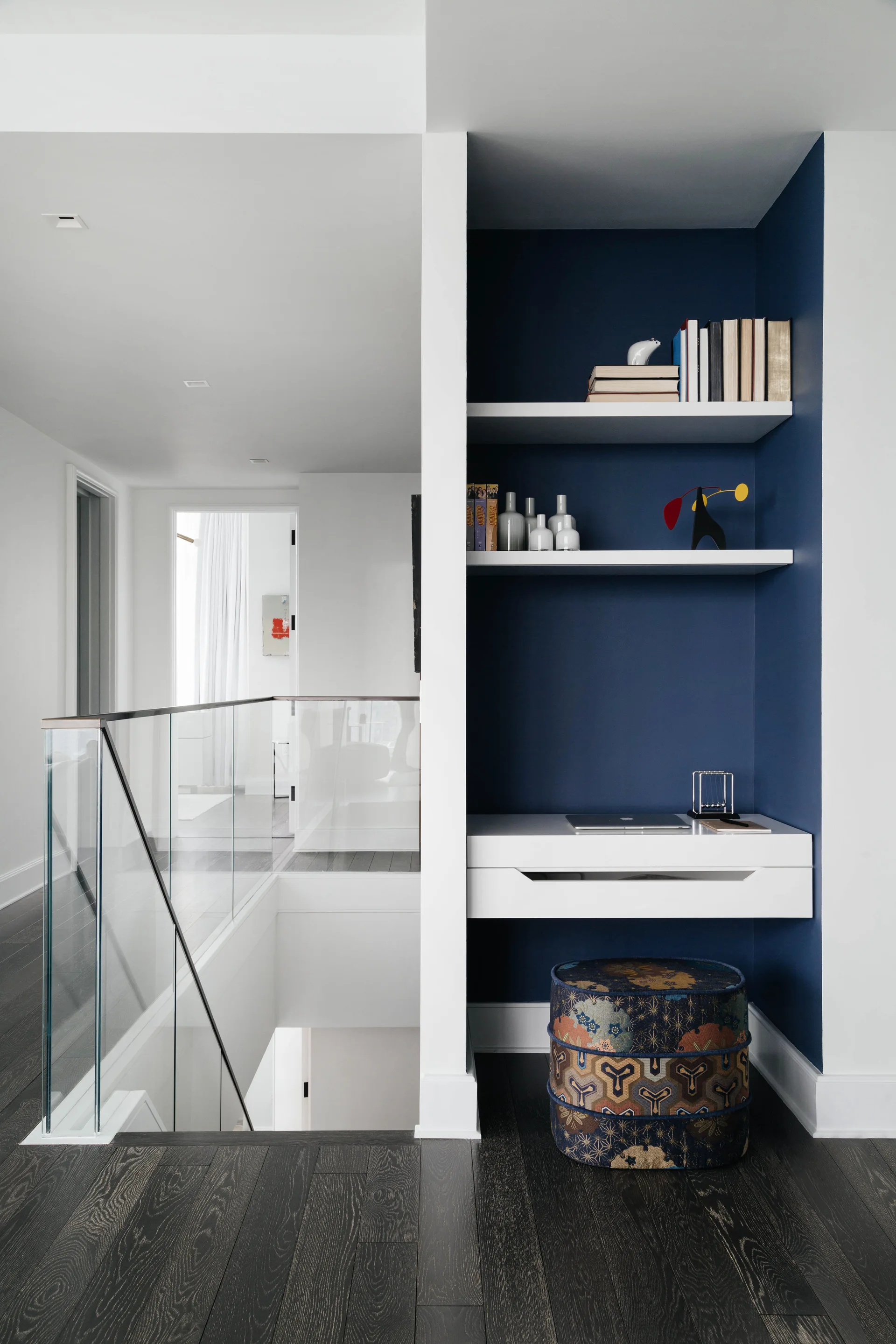 A recessed navy blue wall alcove features a white floating desk and patterned ottoman next to a staircase with glass railings.