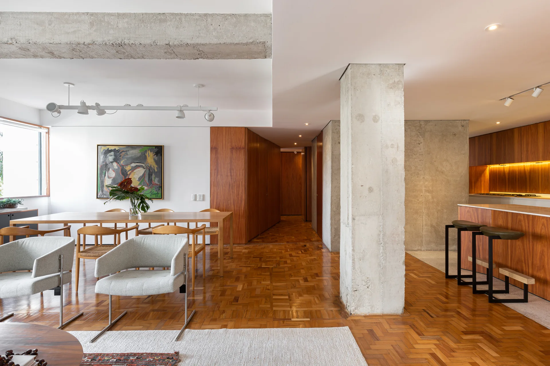 An open-plan dining space features a wooden table, grey armchairs, a prominent raw concrete column, and warm wood-paneled walls.