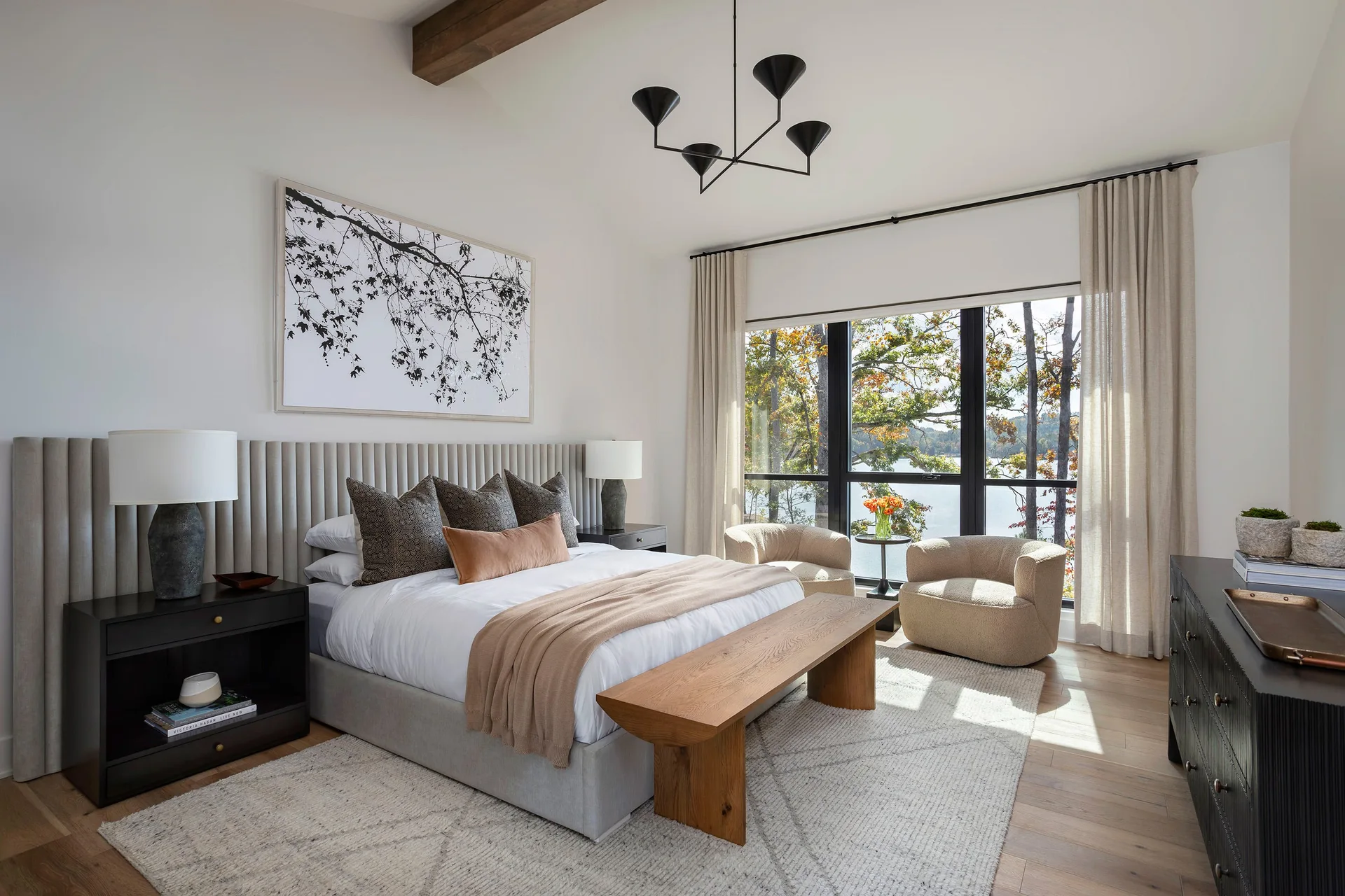An organic modern bedroom features a bed with a channel-tufted headboard wall, a wood bench, two curved armchairs, and a large window showing forest views.
