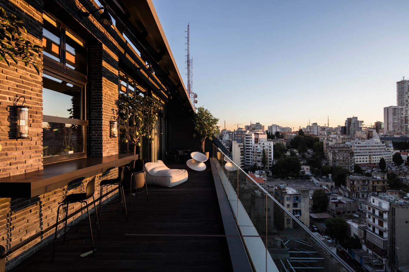 An urban rooftop terrace features a dark brick wall with a bar counter, black stools, and a glass railing overlooking a city skyline.