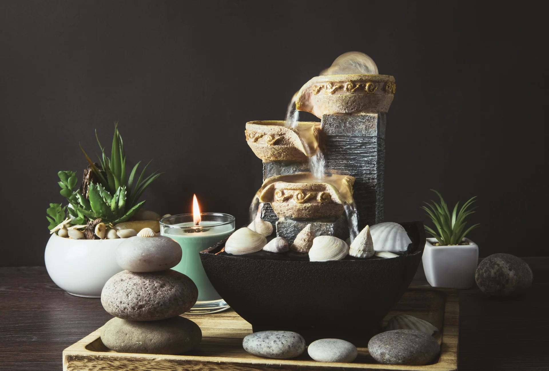 A tiered tabletop water fountain sits on a wooden tray alongside stacked stones, seashells, potted succulents, and a lit green candle against a dark background.
