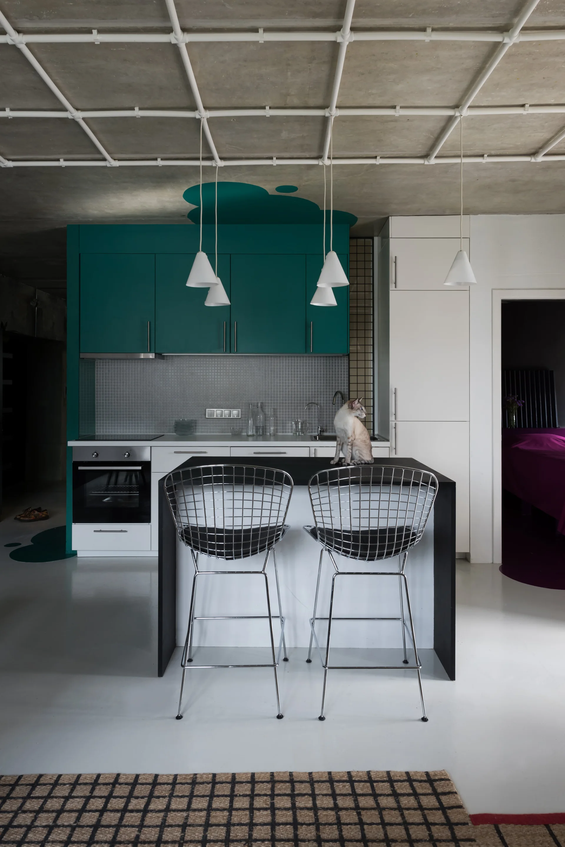 A modern kitchen featuring teal and white cabinets, a black-topped island with wire-frame bar stools, and a white pipe grid on a concrete ceiling.