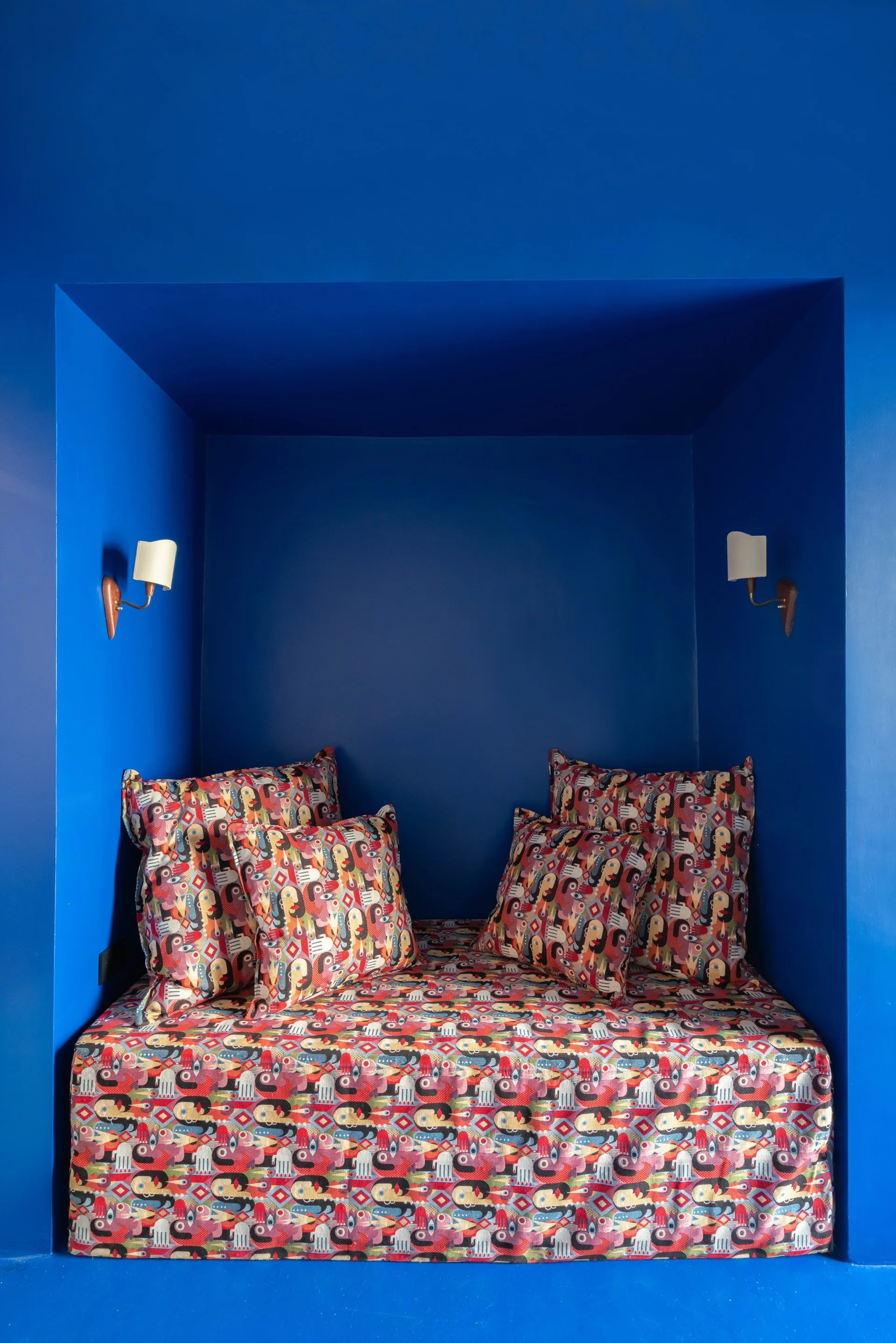 A recessed daybed nook painted entirely in deep cobalt blue features a mattress and matching pillows covered in a colorful patterned fabric.