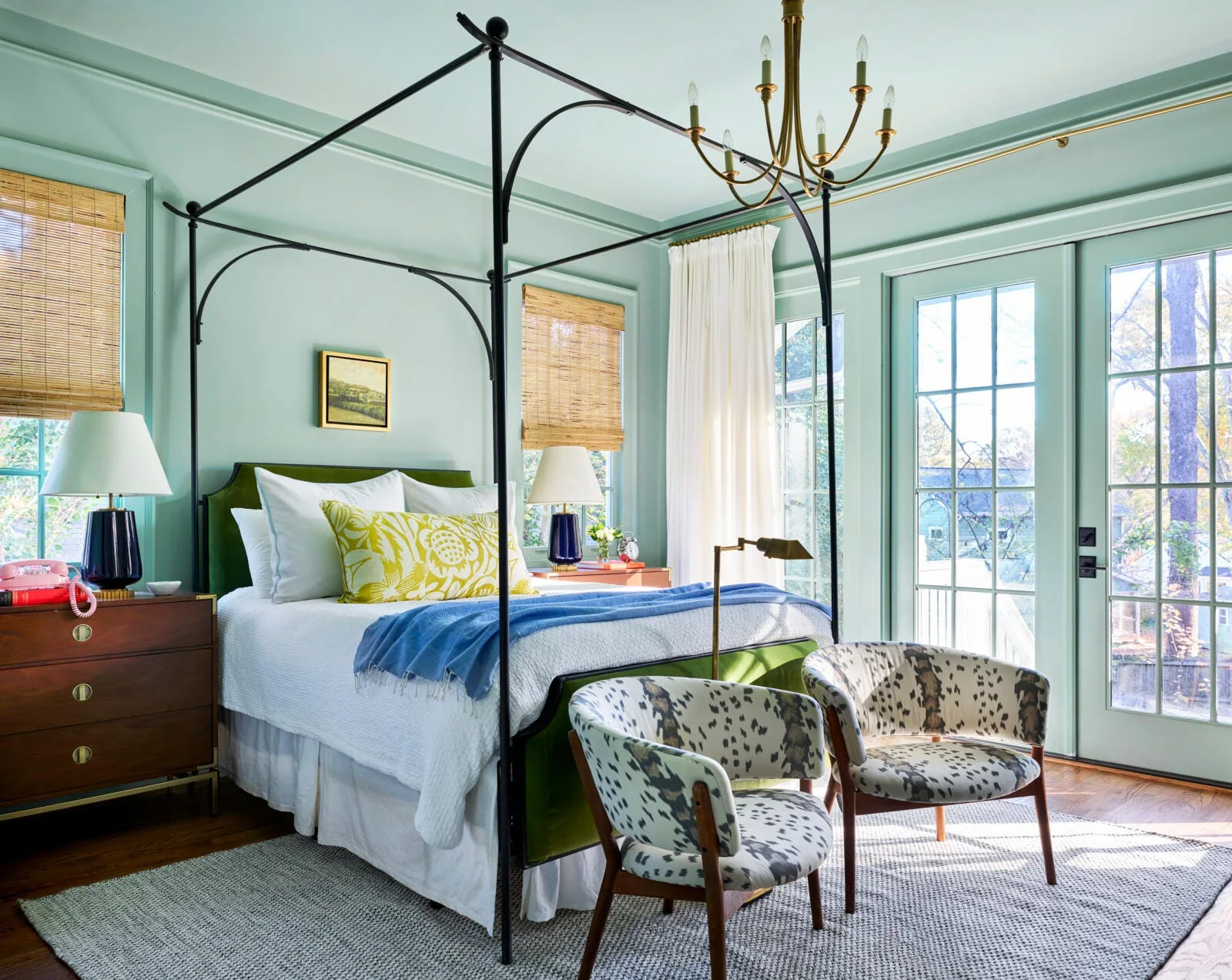 A mint green bedroom features a black metal canopy bed, wooden nightstands with blue lamps, and two spotted accent chairs on a textured rug.