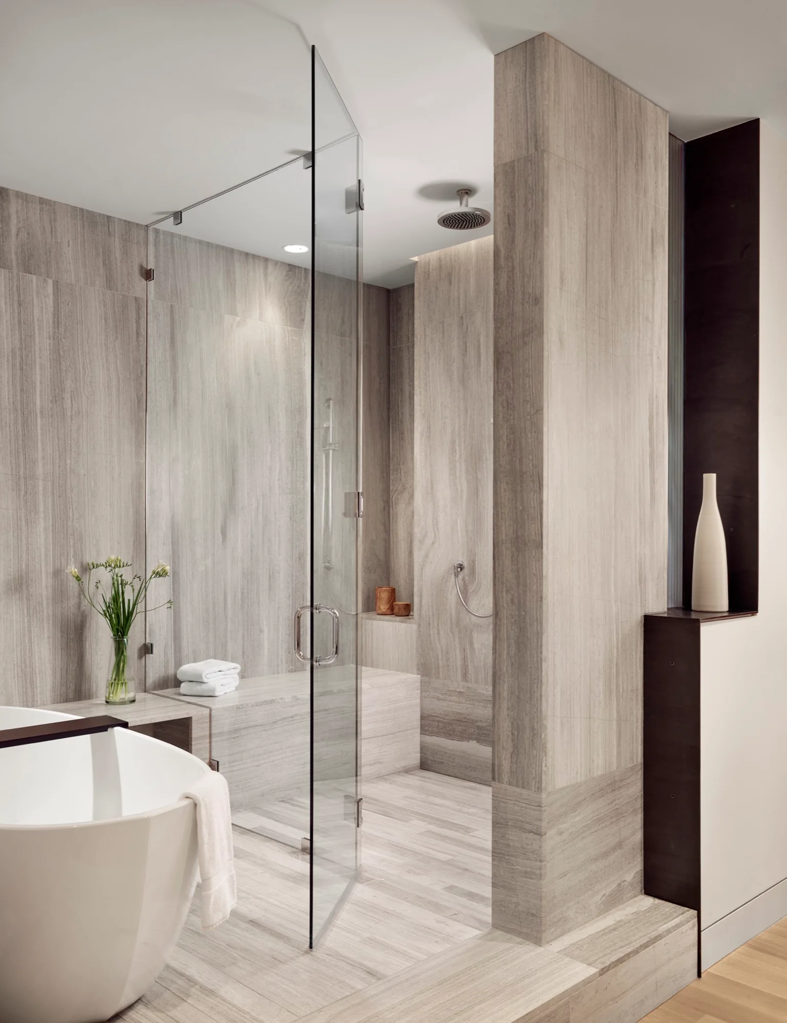 A modern bathroom features a glass-enclosed shower with grey linear-grain tile, a built-in bench, and a white freestanding tub.