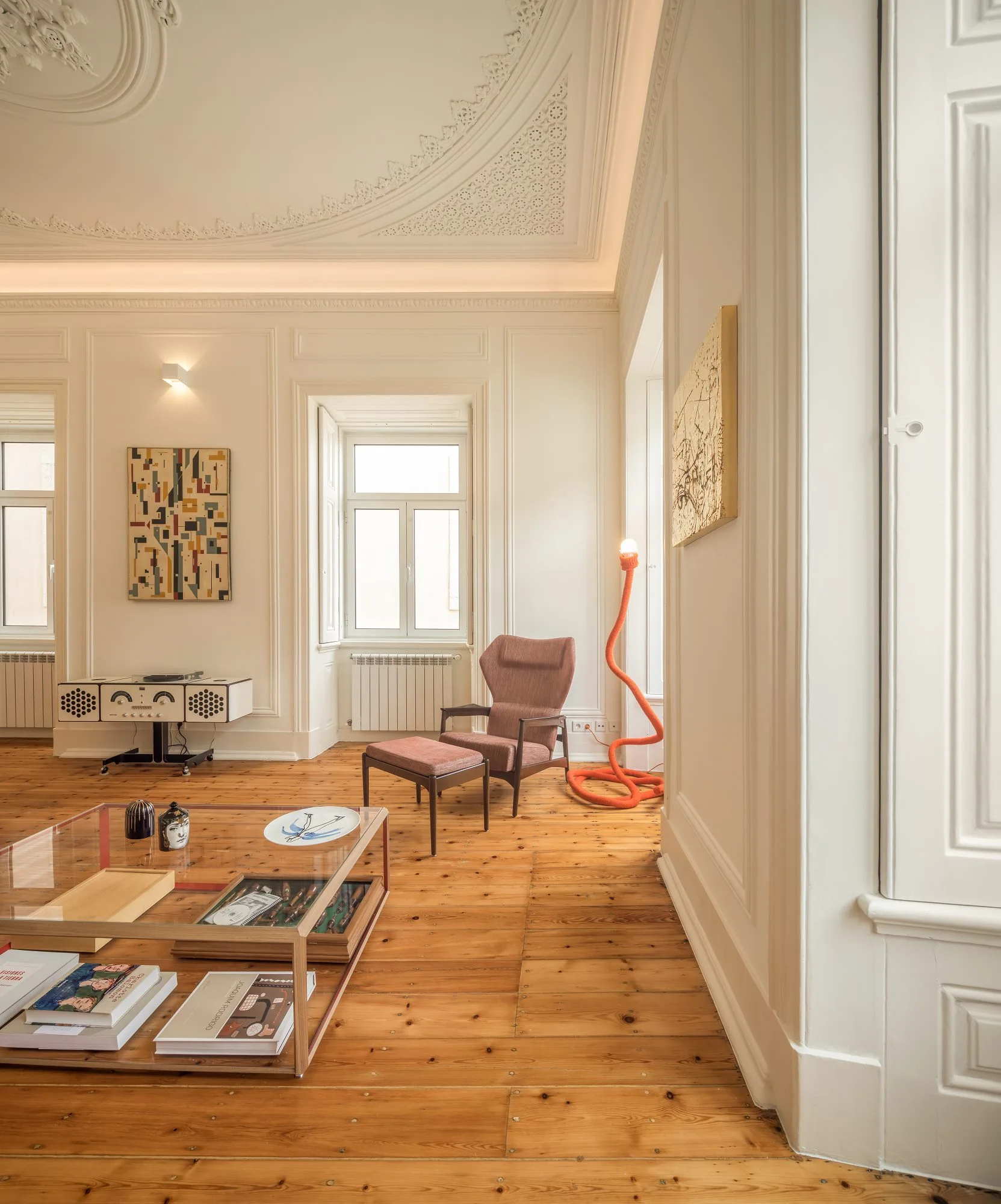 A living room with ornate ceiling moldings, wide plank wood flooring, a pink armchair with matching ottoman, and a glass-topped coffee table.