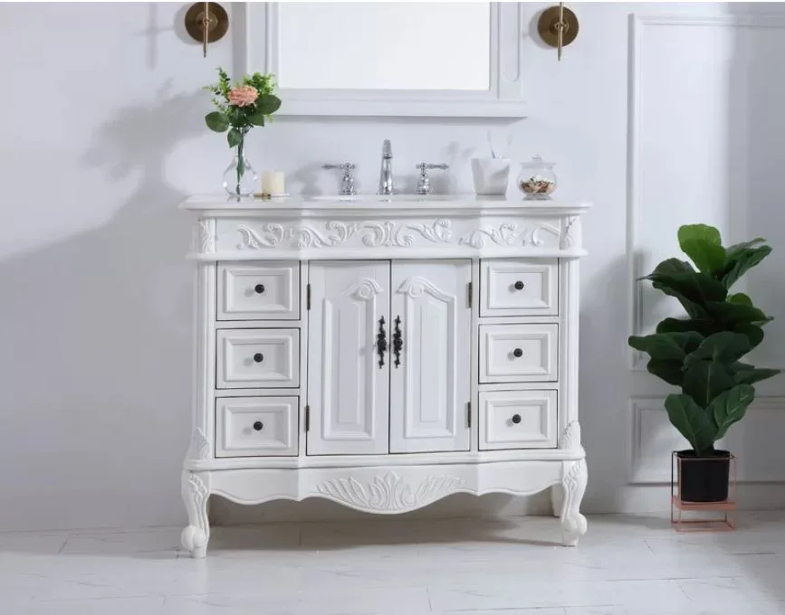 An ornate white bathroom vanity features traditional wood carvings, a stone countertop, and chrome fixtures set against a paneled wall with brass sconces.