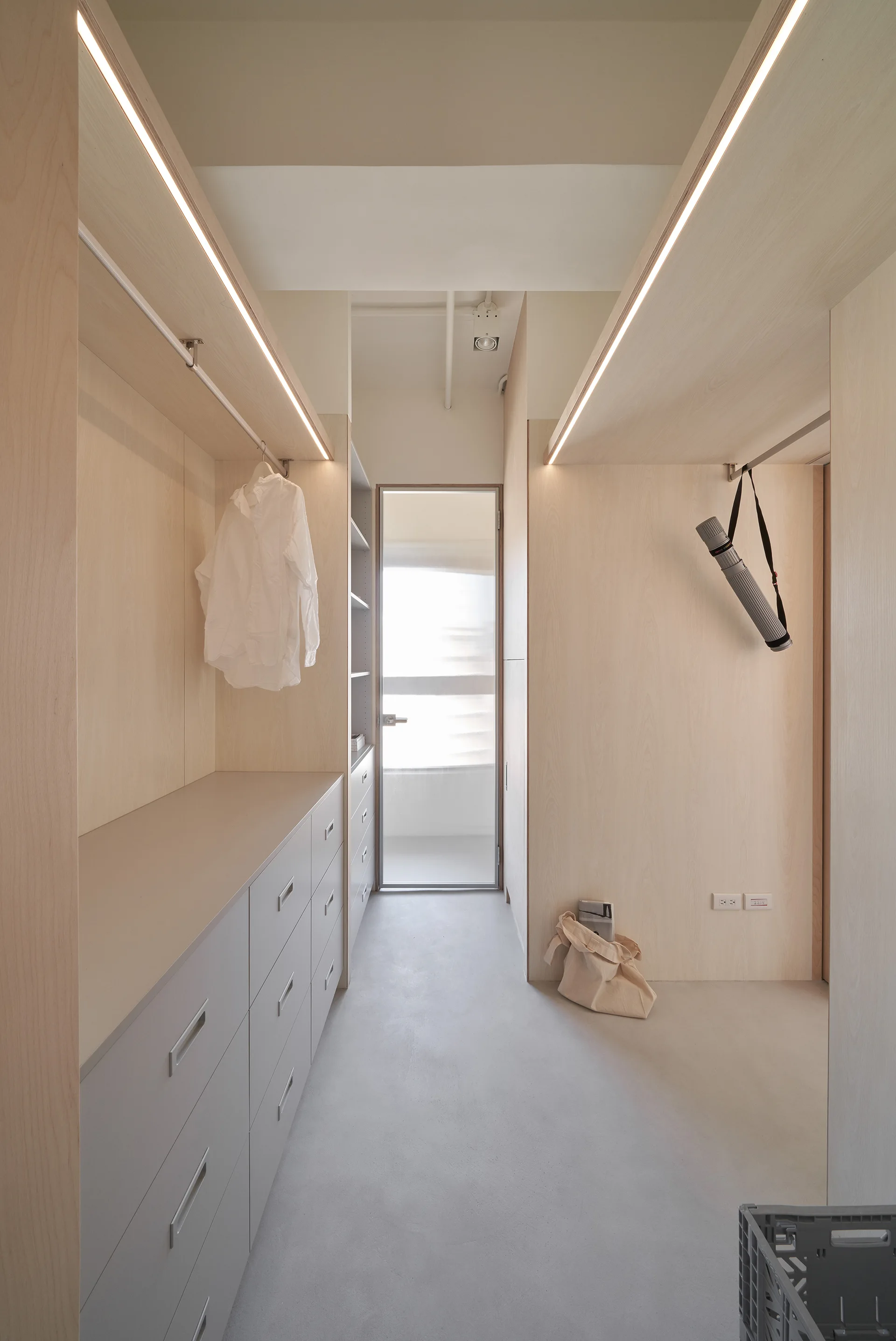 A minimalist walk-in closet featuring light wood cabinetry, built-in drawer units, a concrete floor, and integrated LED strip lighting.