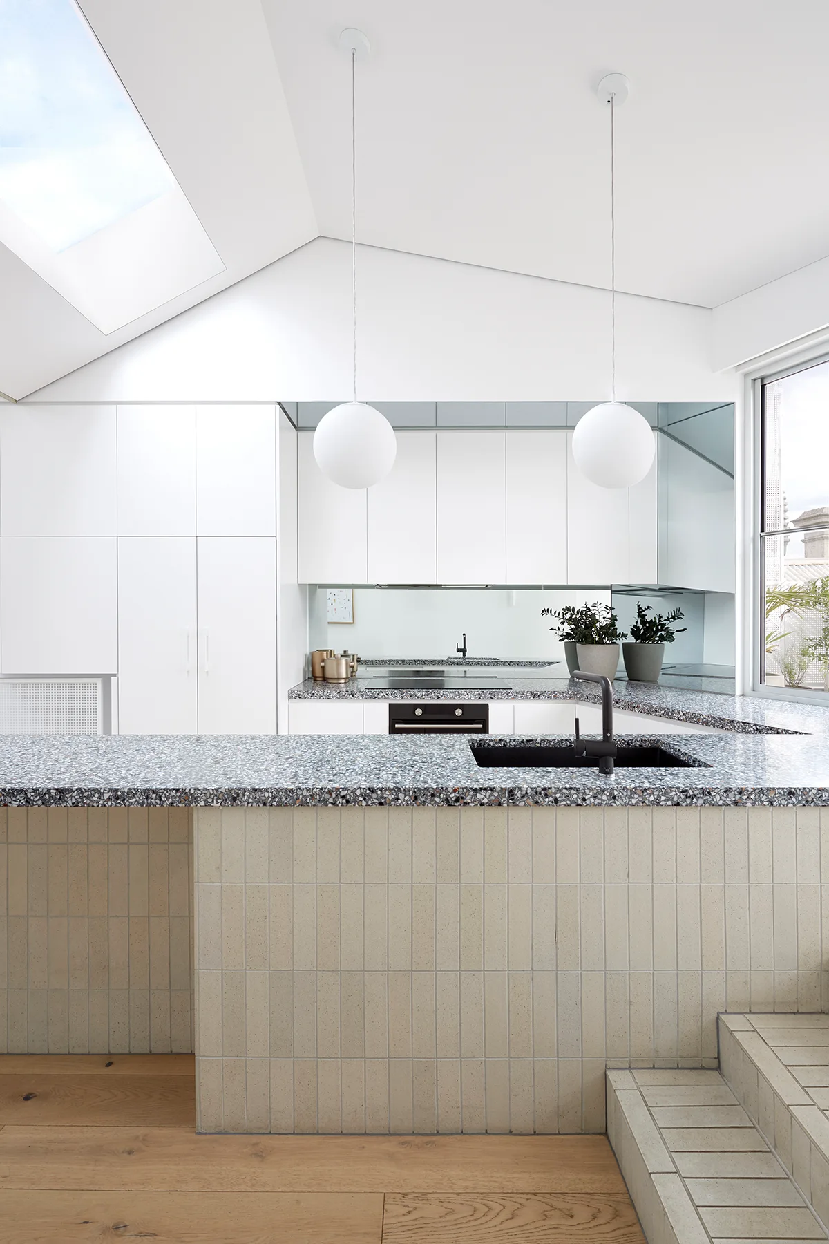 A modern white kitchen features a large island with a speckled terrazzo countertop, a vertically tiled beige base, and spherical white pendant lights.