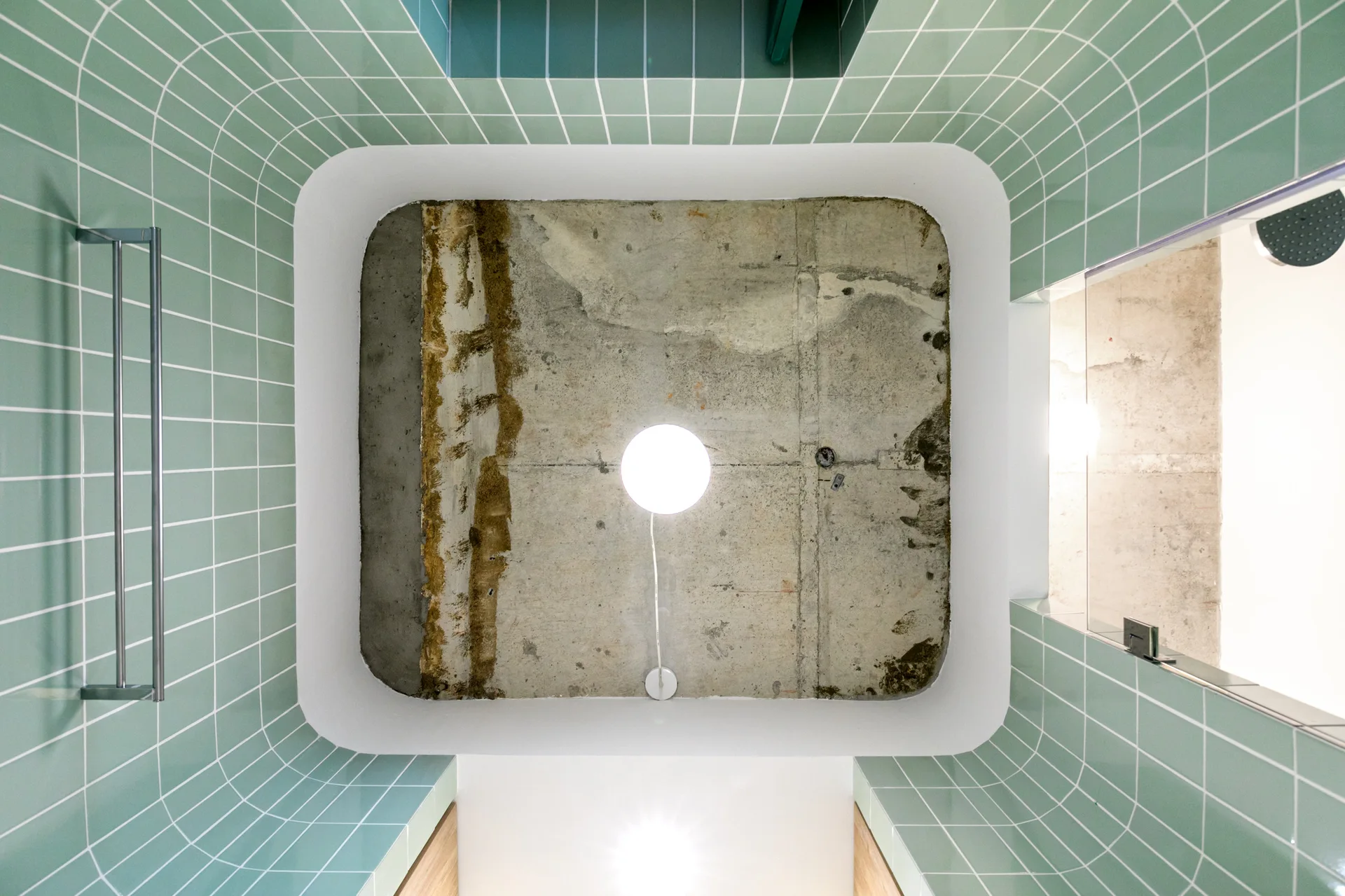 A low-angle view of a bathroom showing light green square-tiled walls curving up to a raw concrete ceiling with a glowing globe pendant light.