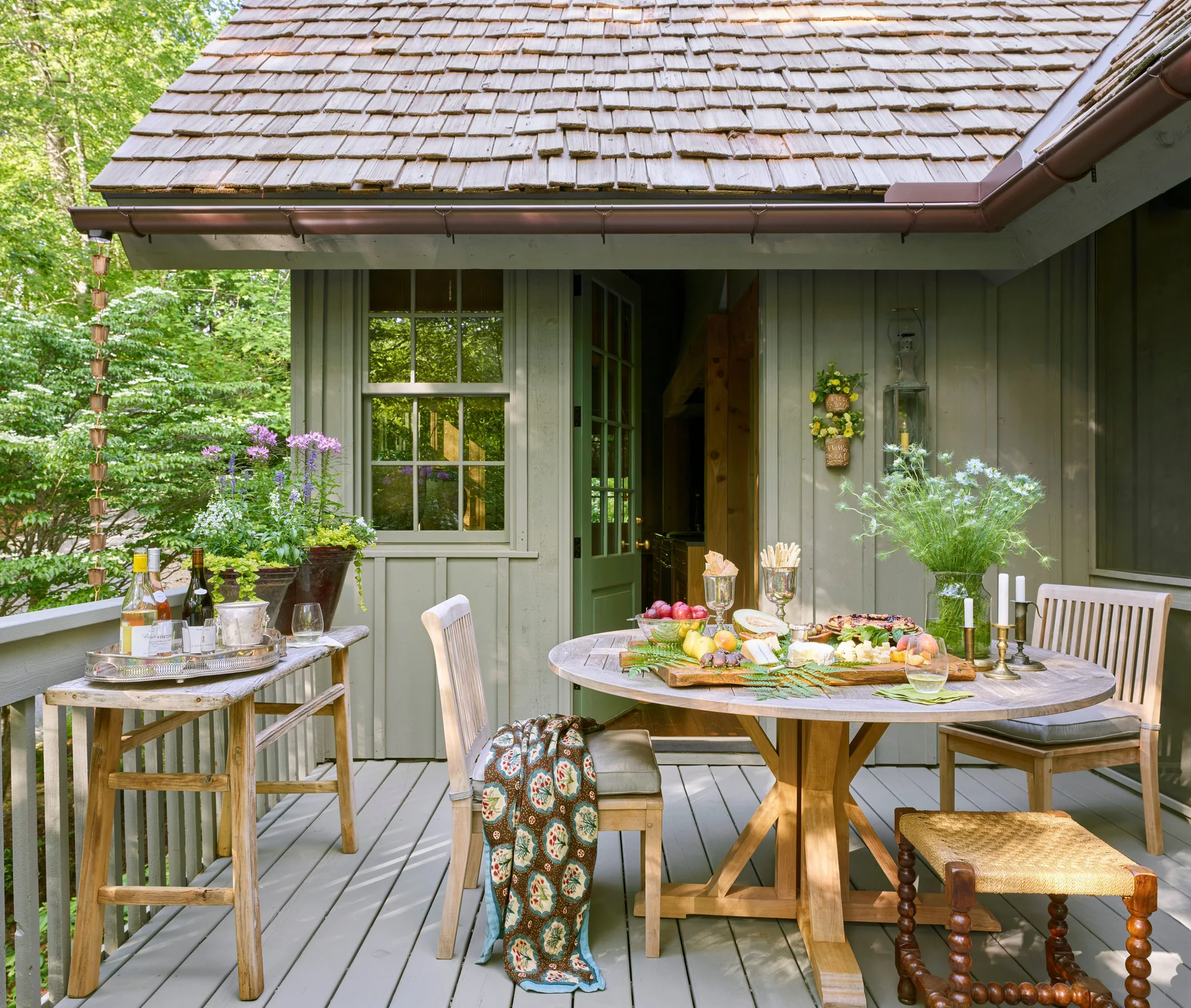 A circular wooden dining table with a food spread on a grey deck next to a house with sage green siding and a cedar shake roof.