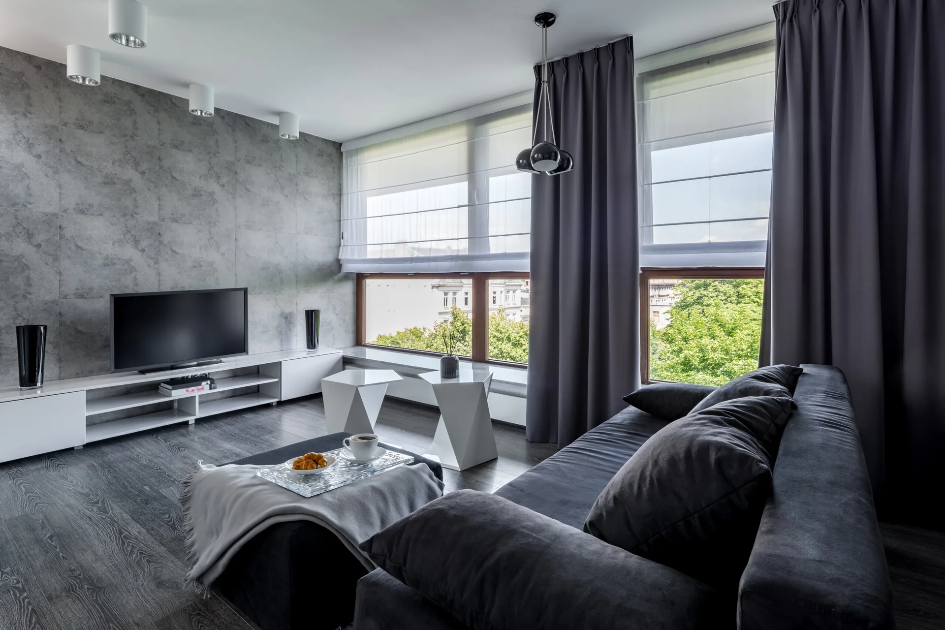 A modern living room features a dark grey sofa, white geometric side tables, a low-profile TV console, and a grey concrete-effect accent wall.