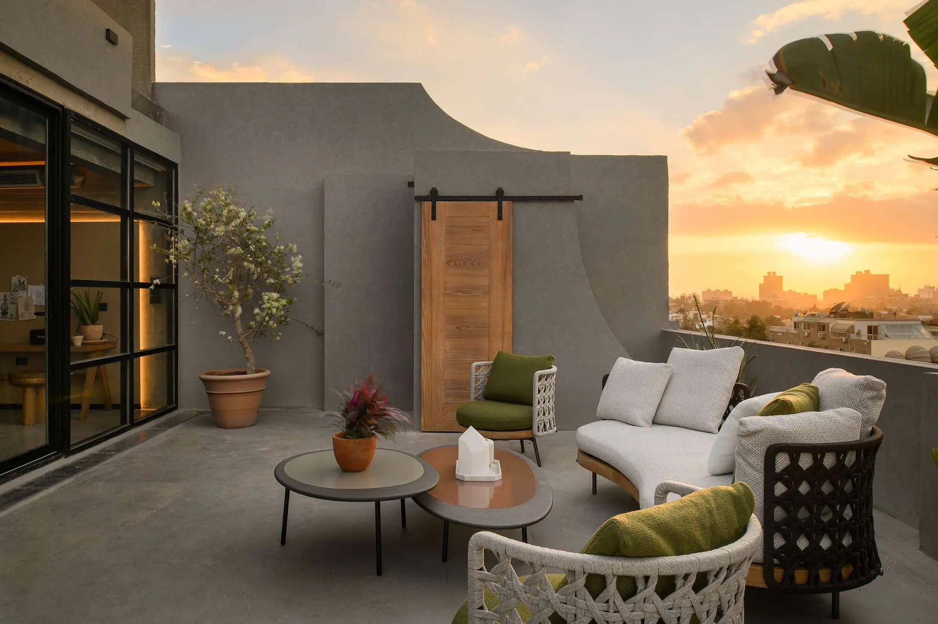 A modern gray rooftop terrace features a curved white sofa, woven armchairs with green cushions, nesting coffee tables, and a wooden sliding barn door.