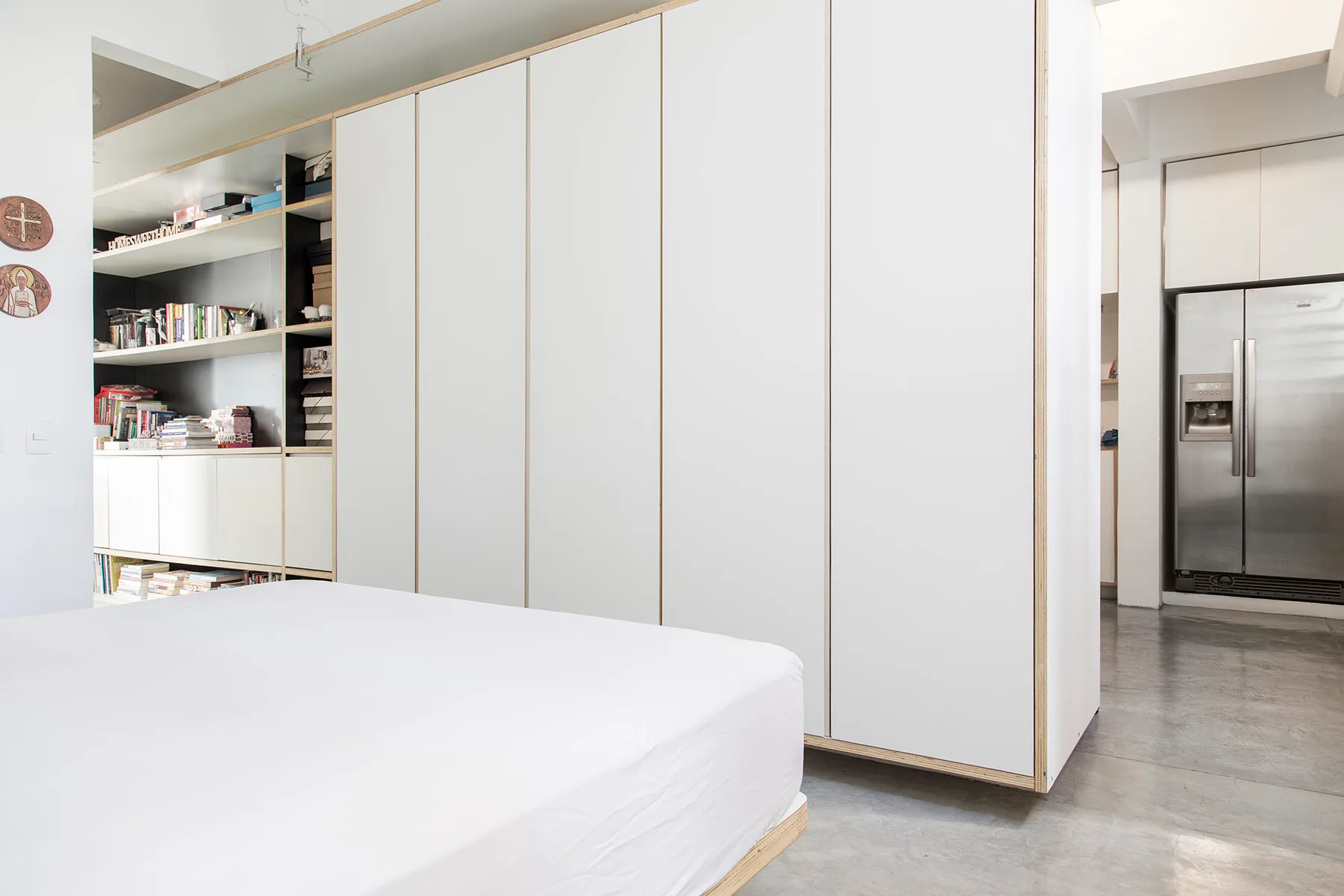 A minimalist bedroom featuring a white bed next to floor-to-ceiling cabinetry with exposed plywood edges on a polished concrete floor.