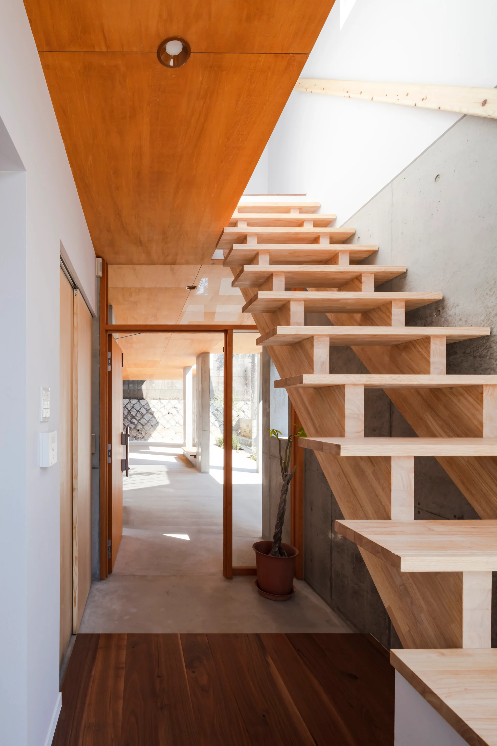 An entryway featuring a wooden open-tread staircase built against a smooth concrete wall under a warm wood-paneled ceiling.