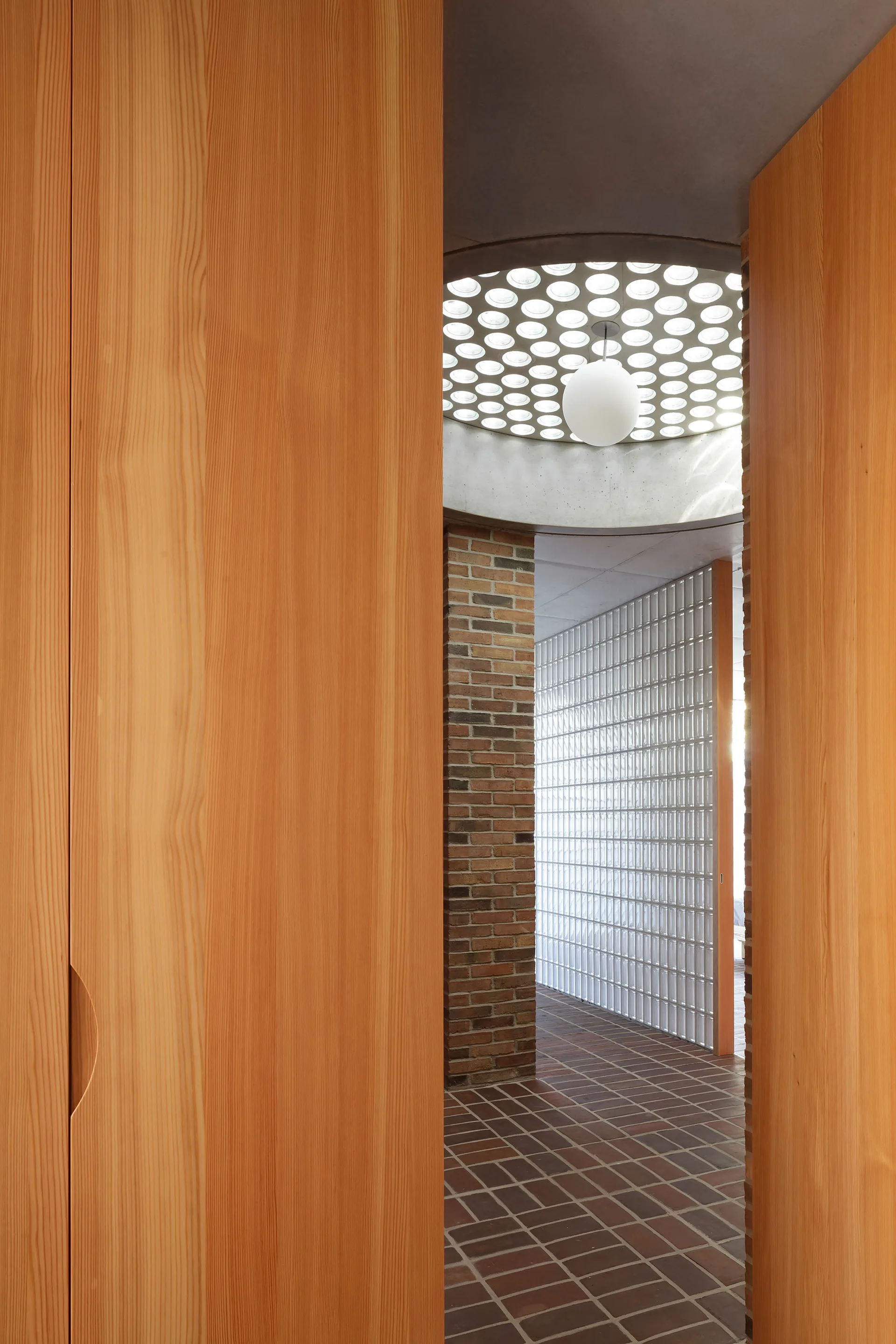 A modernist hallway featuring natural wood cabinetry, an exposed brick wall, a glass block partition, and a circular skylight with a pendant lamp.