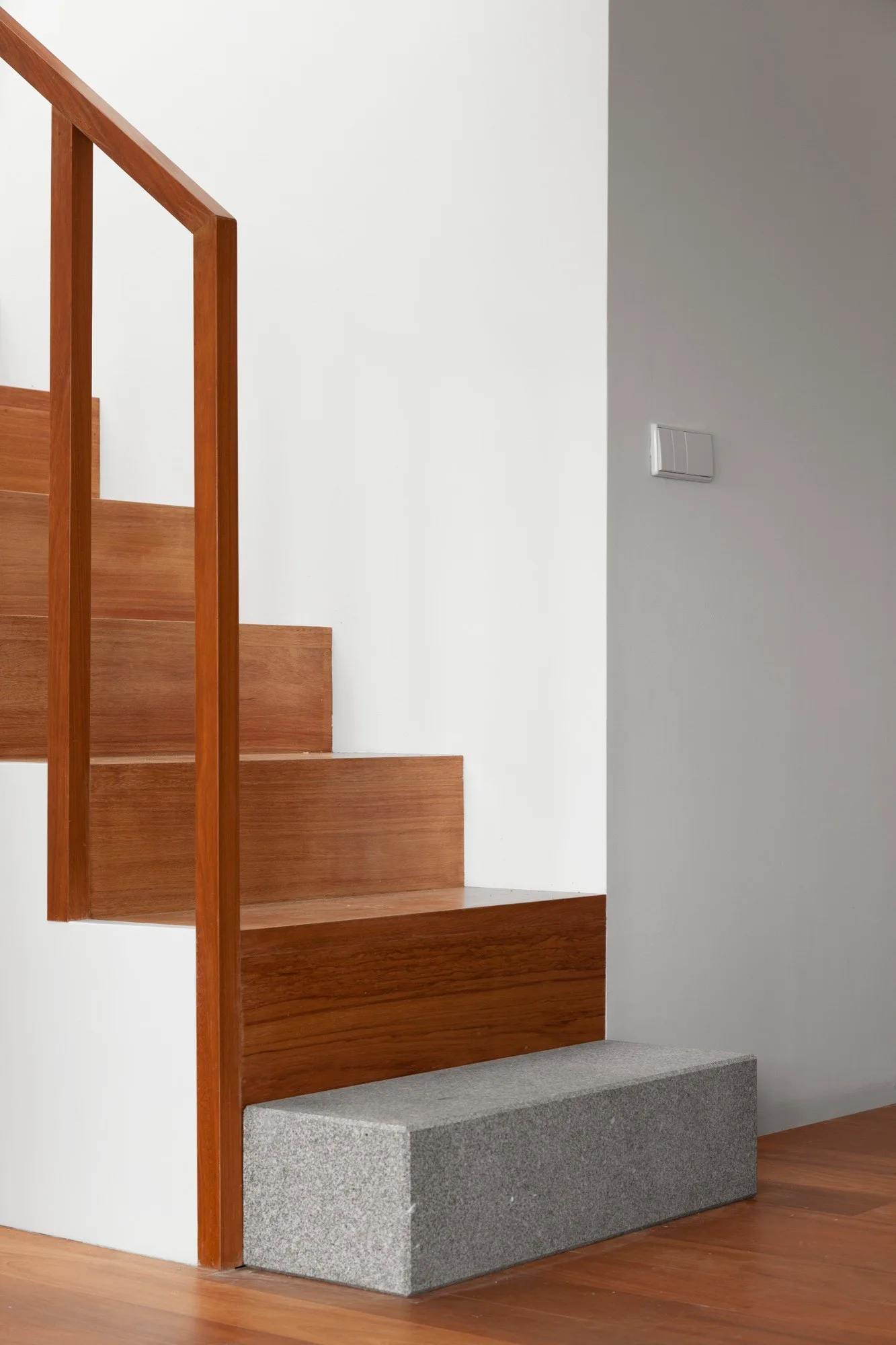 A minimalist wooden staircase with warm timber treads, a matching banister, and a grey stone block as the bottom step against a white wall.