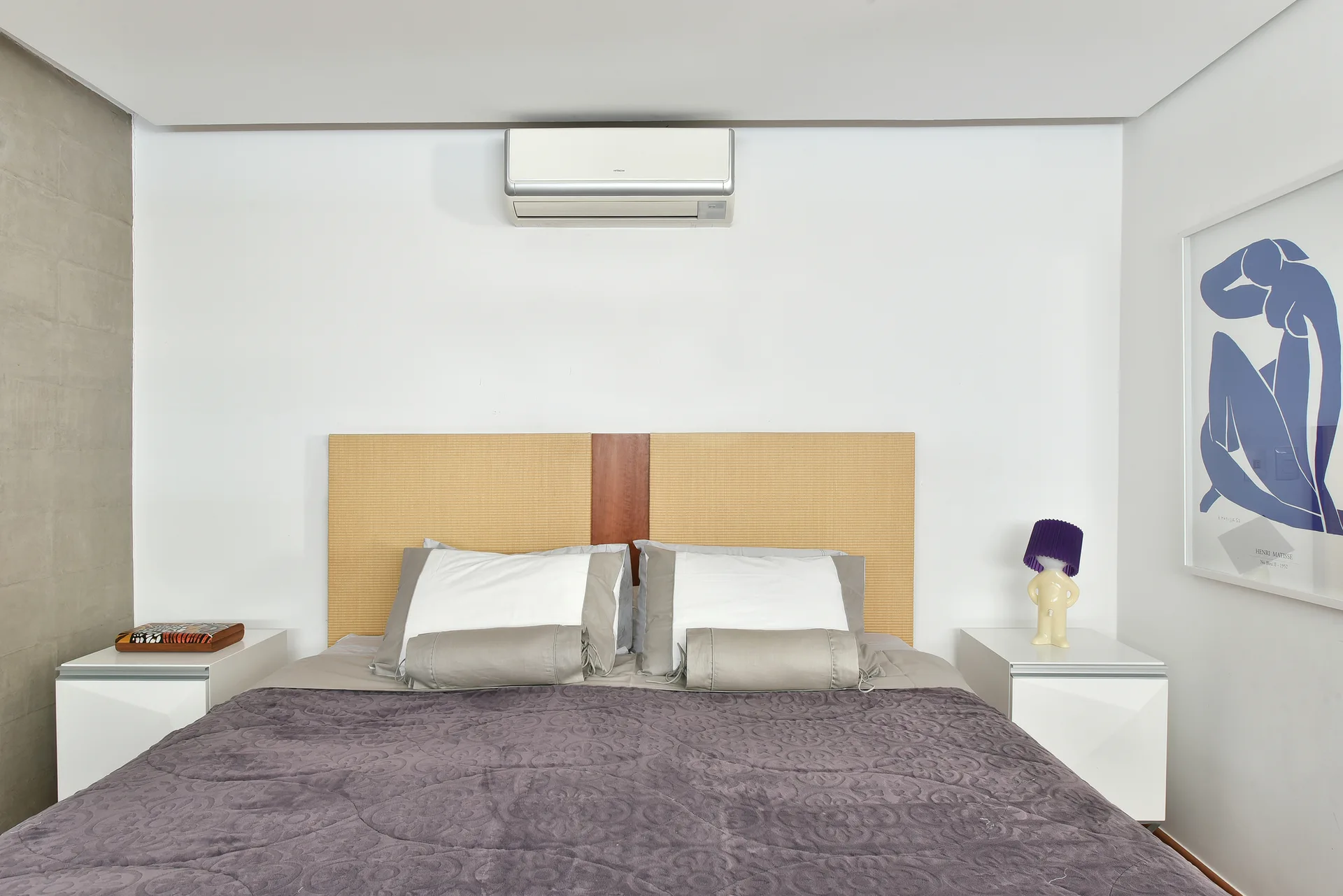 A modern bedroom features a bed with a woven headboard, white nightstands, a purple figure-shaped table lamp, and blue wall art.