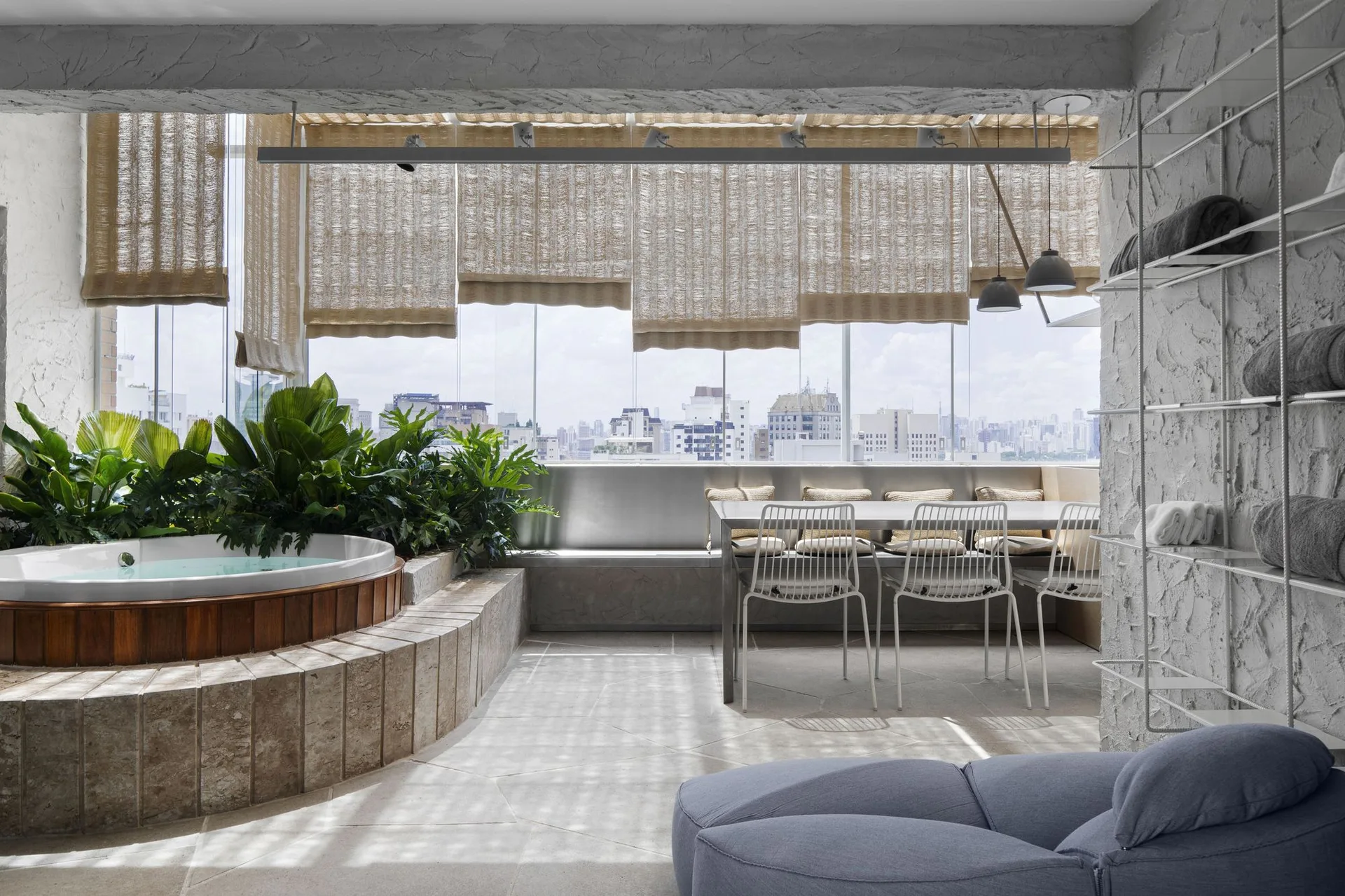 A modern covered terrace featuring a circular wooden hot tub surrounded by plants, a dining area with white wire chairs, and woven Roman shades.