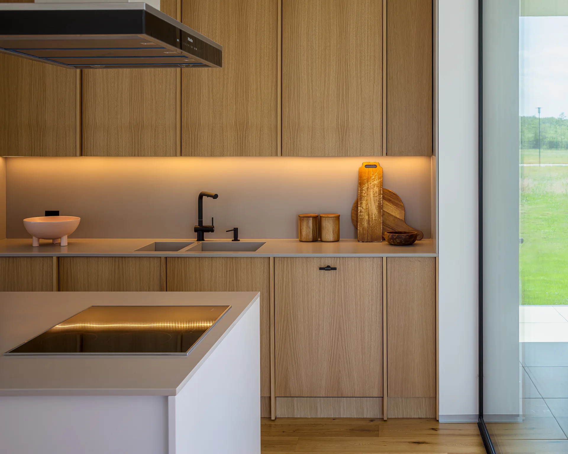 A minimalist kitchen featuring flat-front wood cabinets, a beige countertop with a matte black faucet, and warm under-cabinet strip lighting.