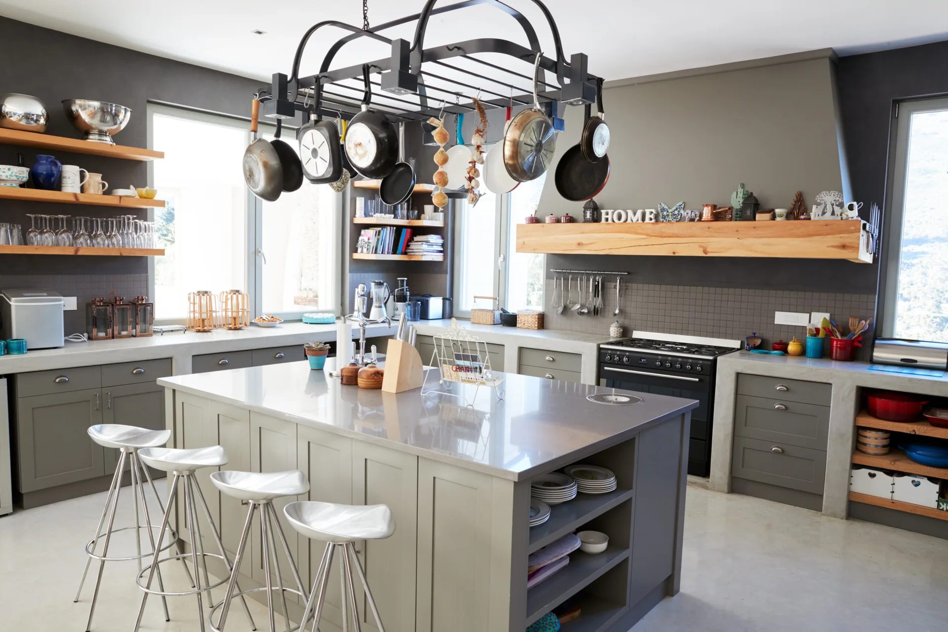 A modern industrial kitchen featuring a grey island with metal bar stools, an overhead black pot rack filled with pans, and wooden floating shelves.