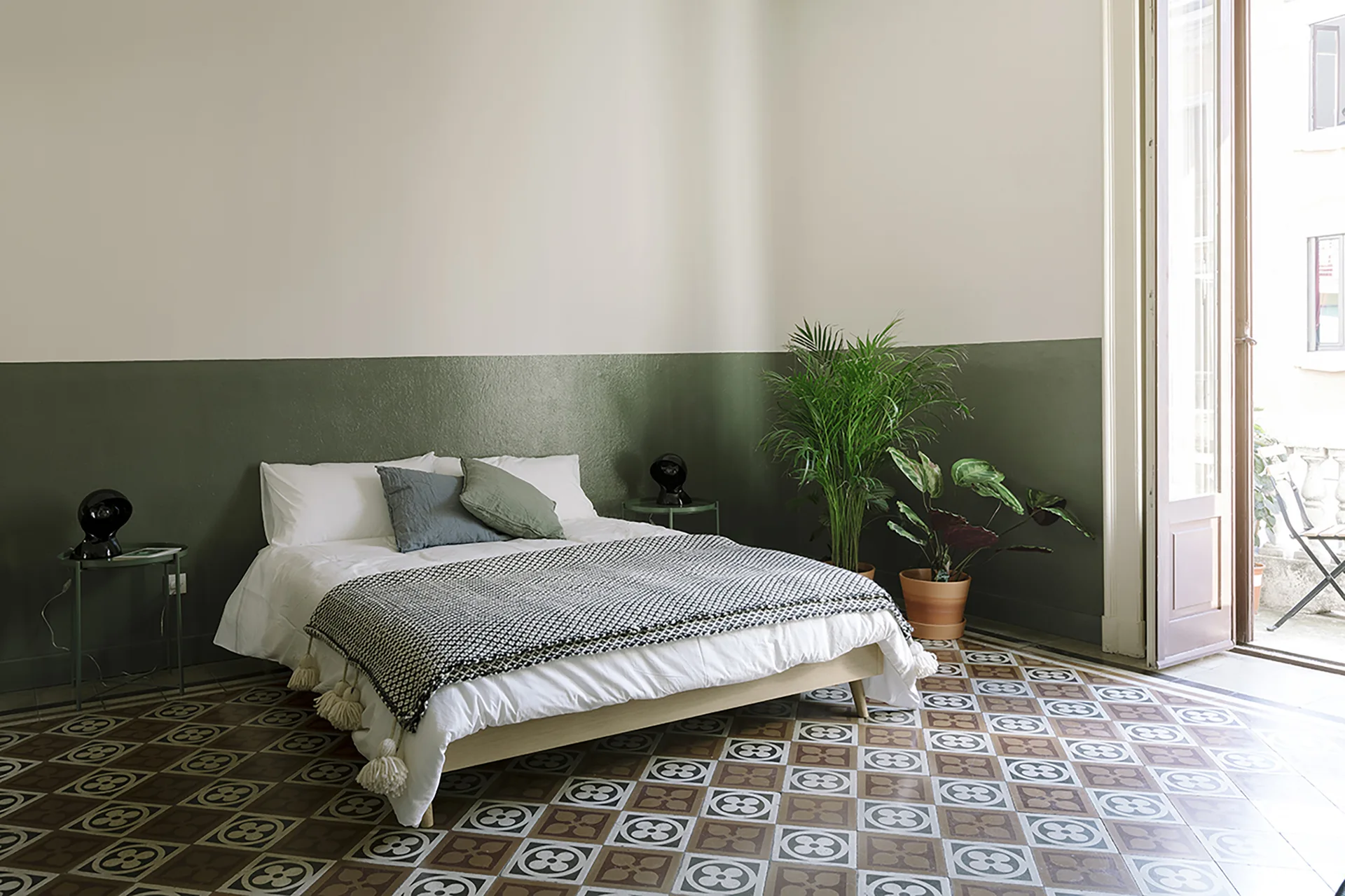 A minimalist bedroom featuring a bed with a tasseled throw blanket, two-tone sage green walls, and a patterned geometric tile floor.