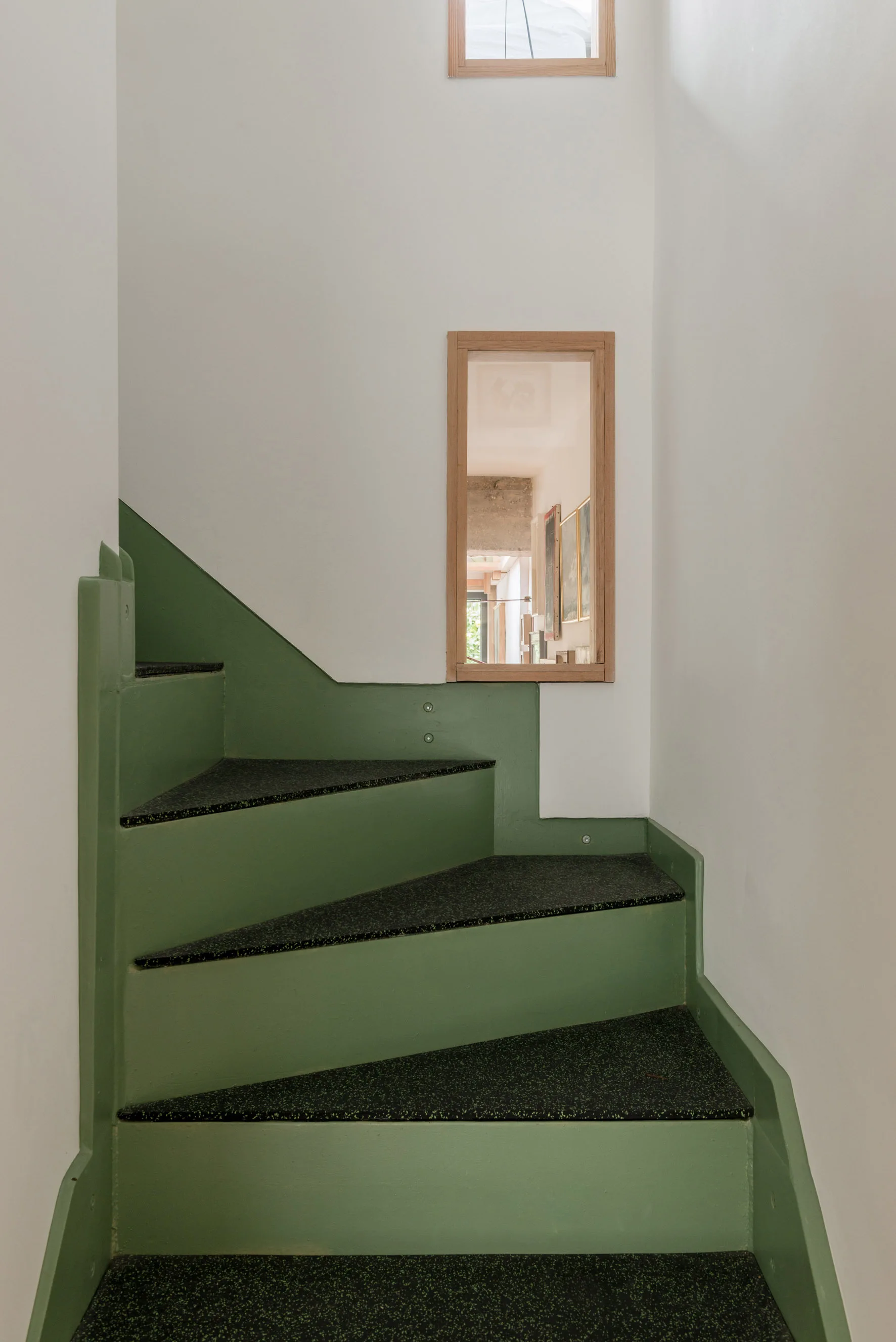 A minimalist staircase with sage green painted risers and dark speckled treads features a wood-framed interior window set into the white wall.