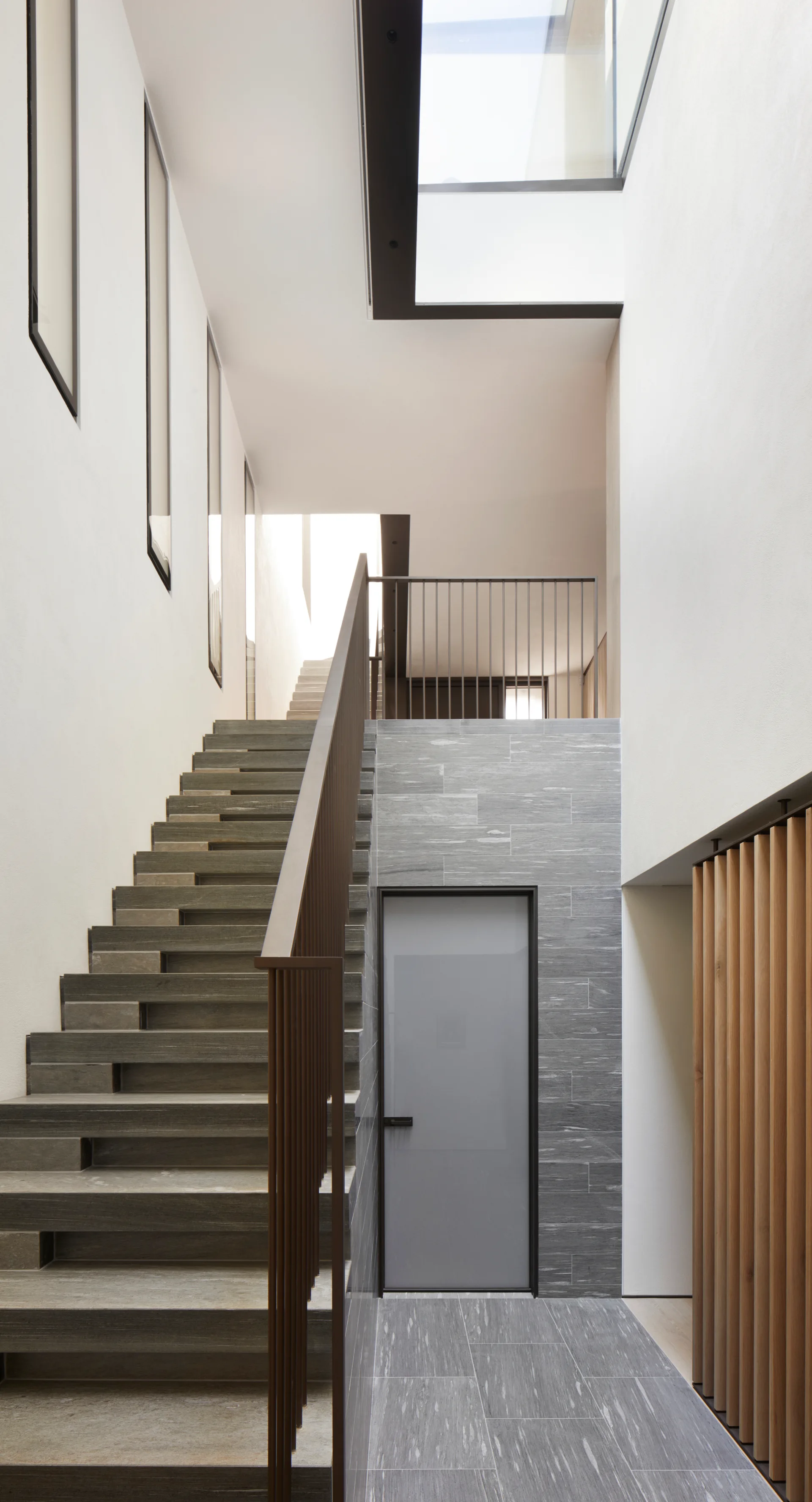 A modern minimalist stairwell features floating grey stone steps, a dark metal railing, an overhead skylight, and a vertical wood slat wall.