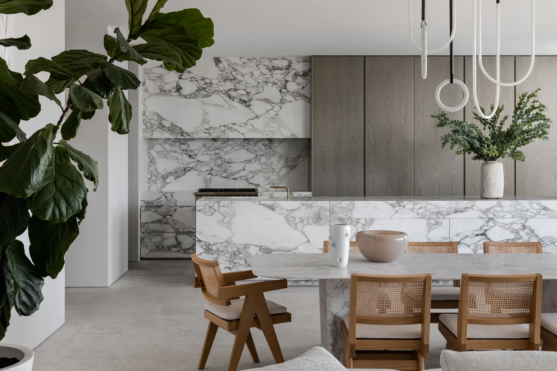 A modern open-plan kitchen and dining room featuring a heavily veined marble waterfall island, matching marble dining table, and wood cabinetry.