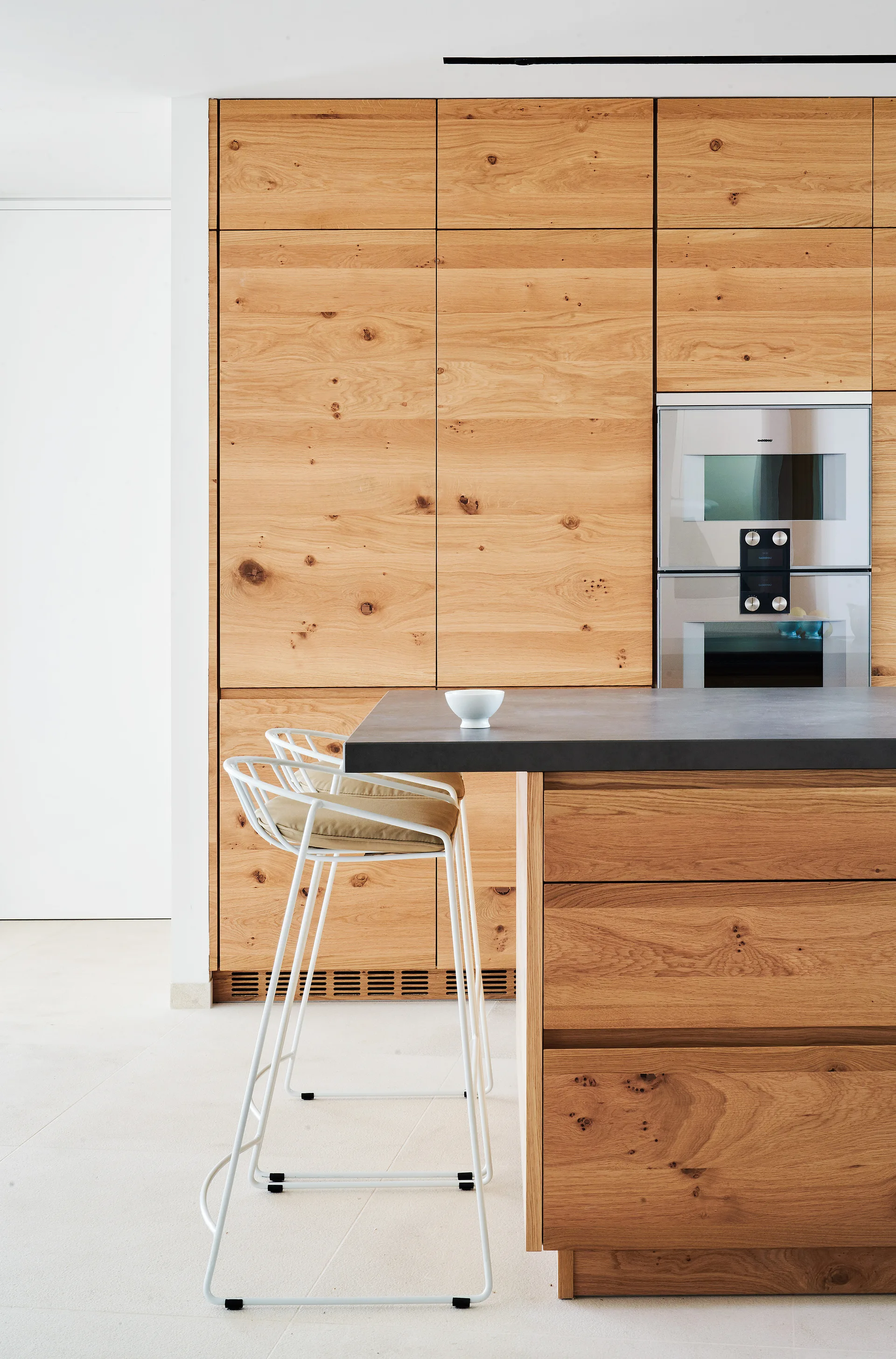 A minimalist kitchen features flat-panel wood cabinets with visible grain, a built-in steel oven, and a dark-topped island with a white bar stool.