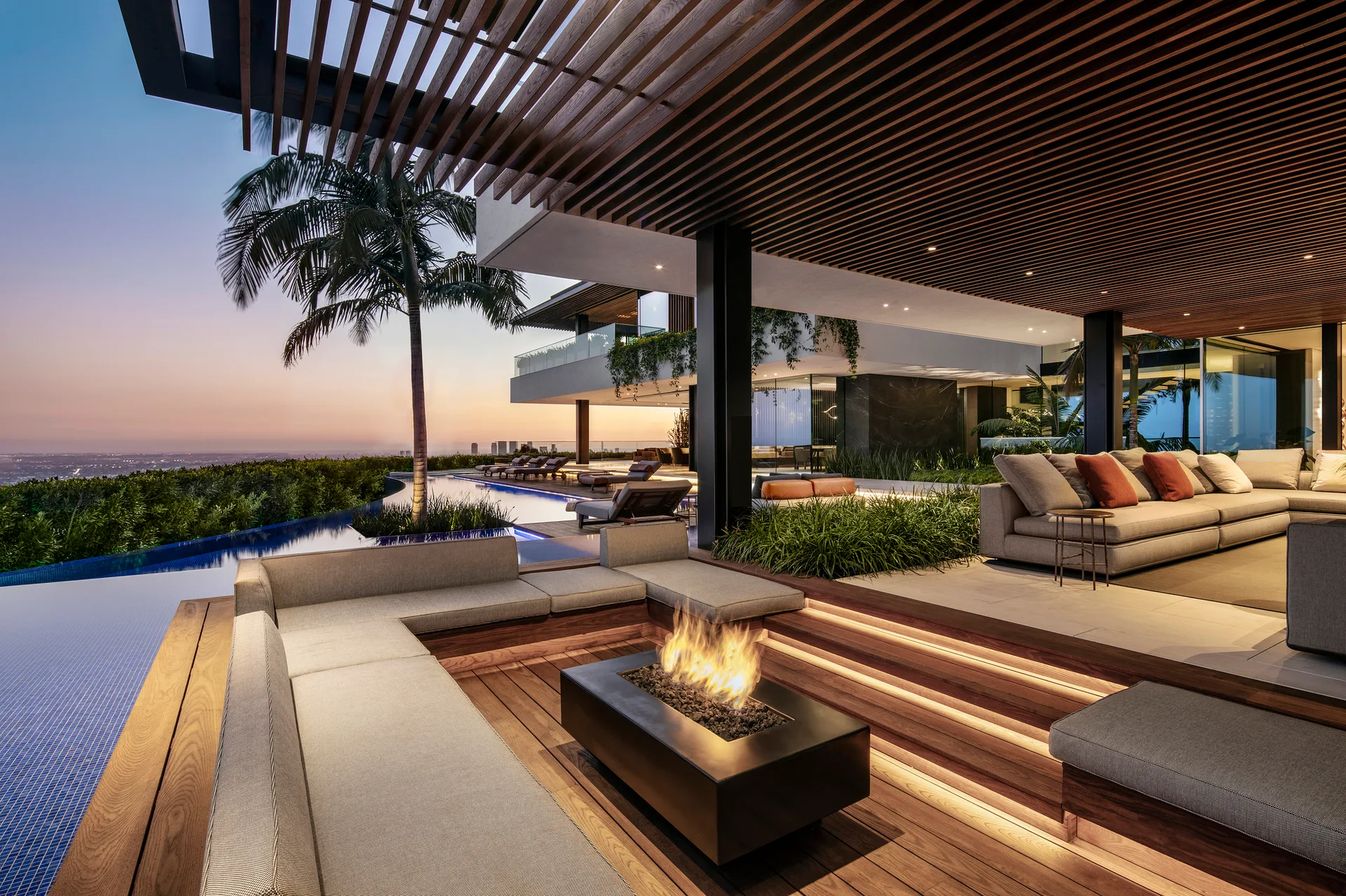 A modern outdoor deck features a sunken seating area with a central fire pit next to an infinity swimming pool under a slatted wooden ceiling.
