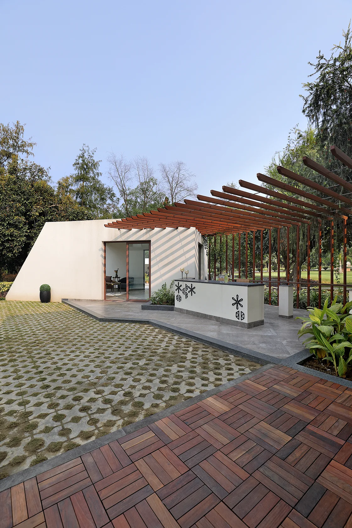 An outdoor patio features a wooden slat pergola shading a white bar counter alongside a tiled walkway, grass grid pavers, and wood decking.