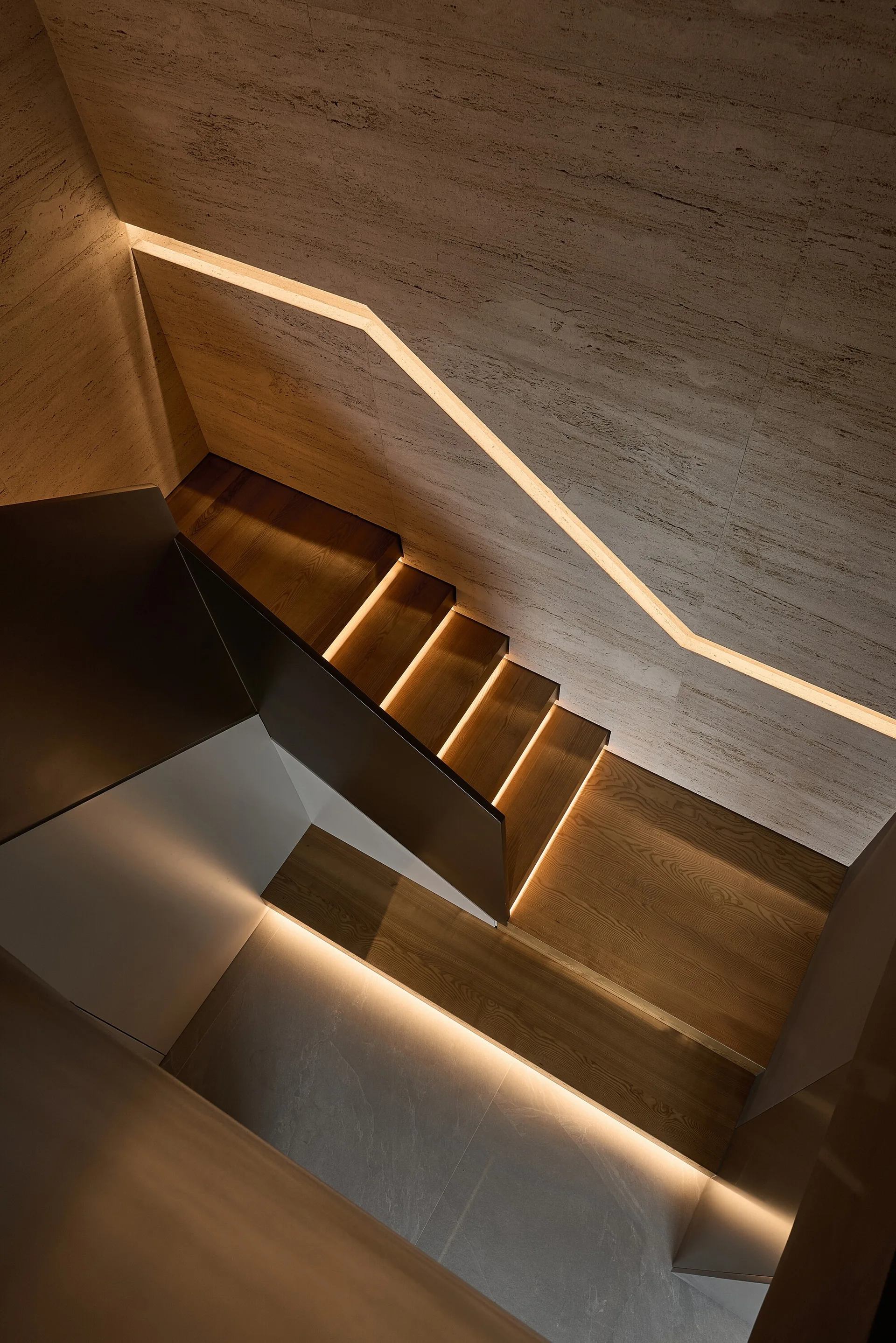 A modern minimalist staircase viewed from above features wooden treads illuminated by warm integrated under-step LED lighting against textured stone walls.