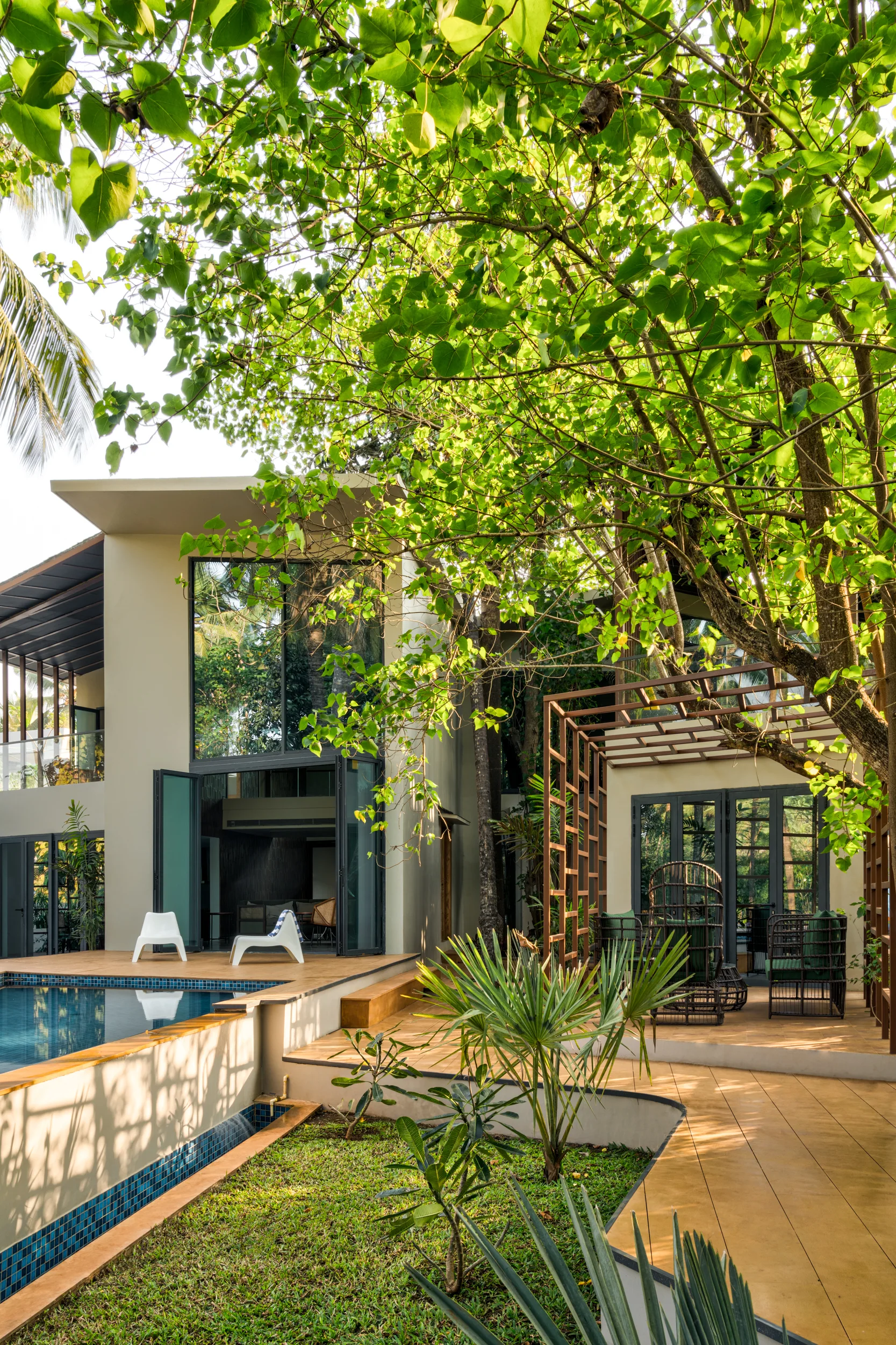 A modern home featuring a swimming pool, light wood decking, and a metal pergola-covered seating area surrounded by trees.