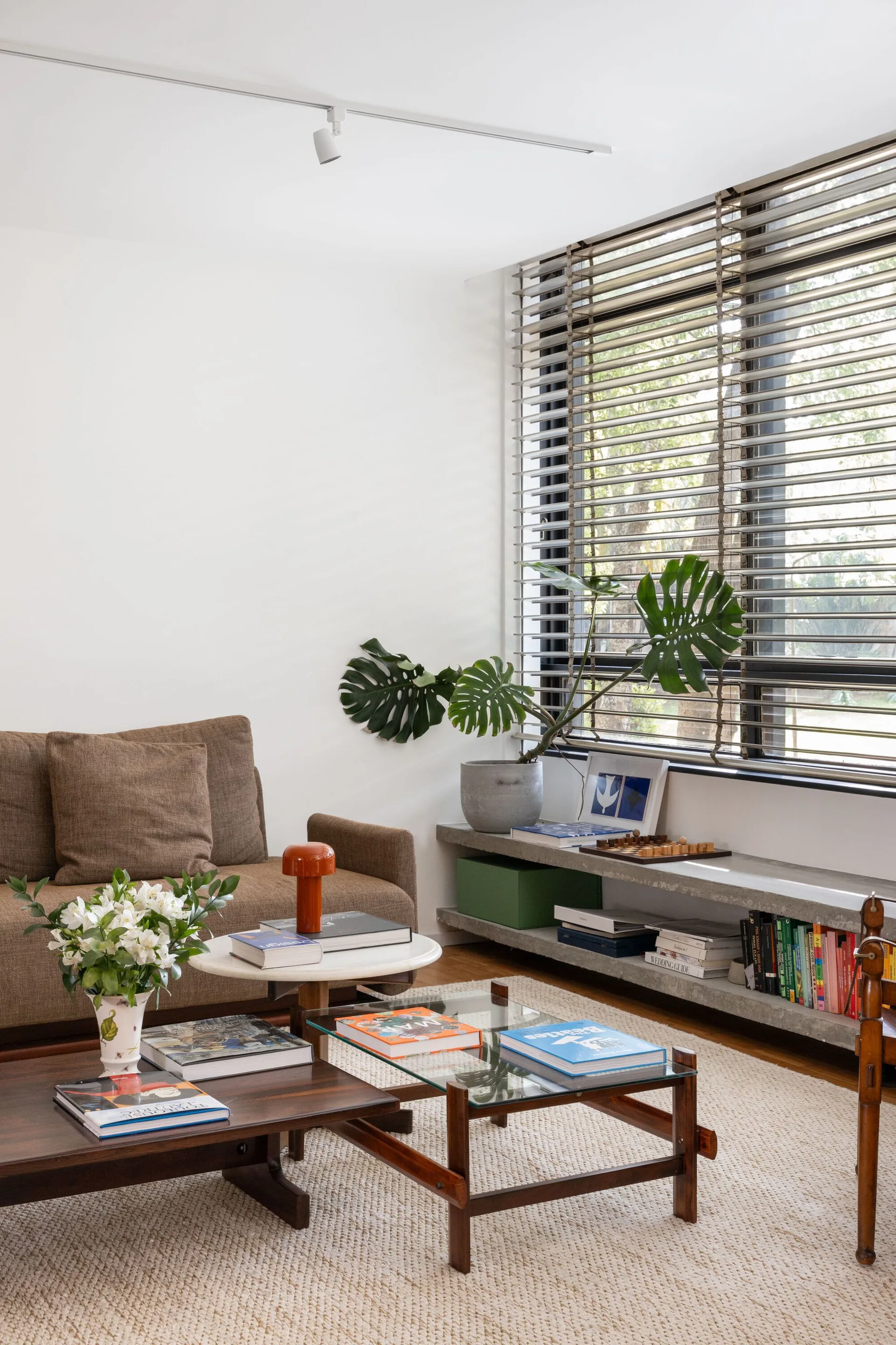 A mid-century modern living room features a brown sofa, tiered coffee tables, a woven rug, and a low concrete ledge shelf under a window.