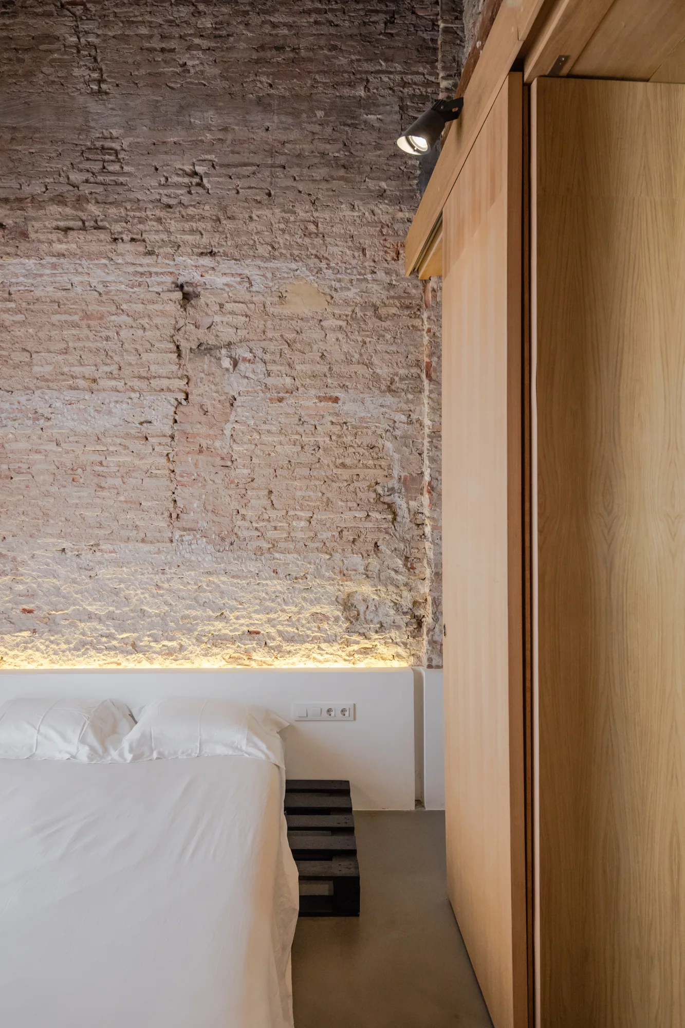 A minimalist bedroom features a white bed against an exposed brick wall highlighted by built-in LED headboard lighting and tall wooden closets.