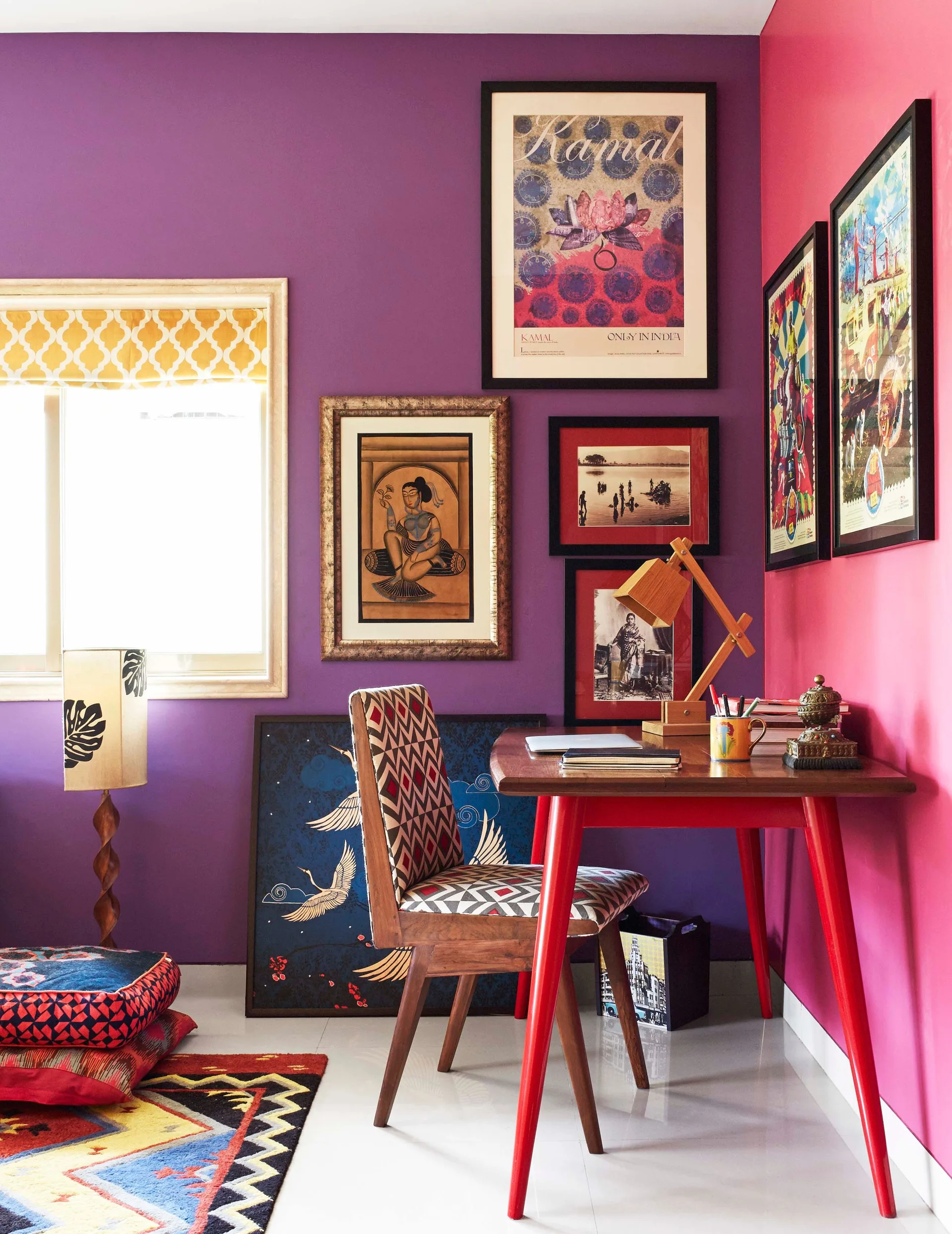 An eclectic home office features a wooden desk with red legs, a patterned chair, and a gallery wall set against purple and pink painted walls.