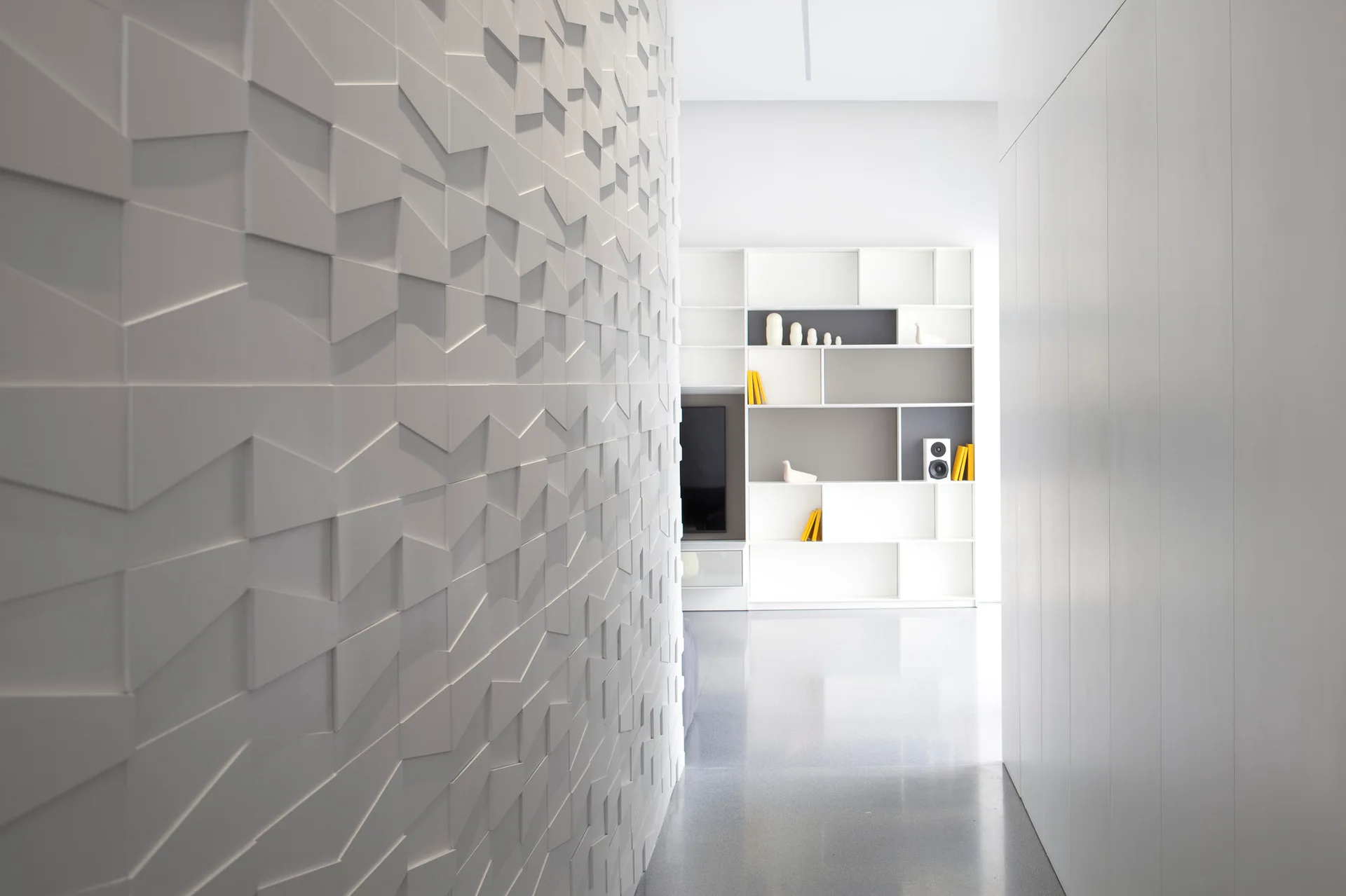 A modern white hallway features a prominent geometric 3D textured wall leading toward a built-in minimalist shelving unit in the background.