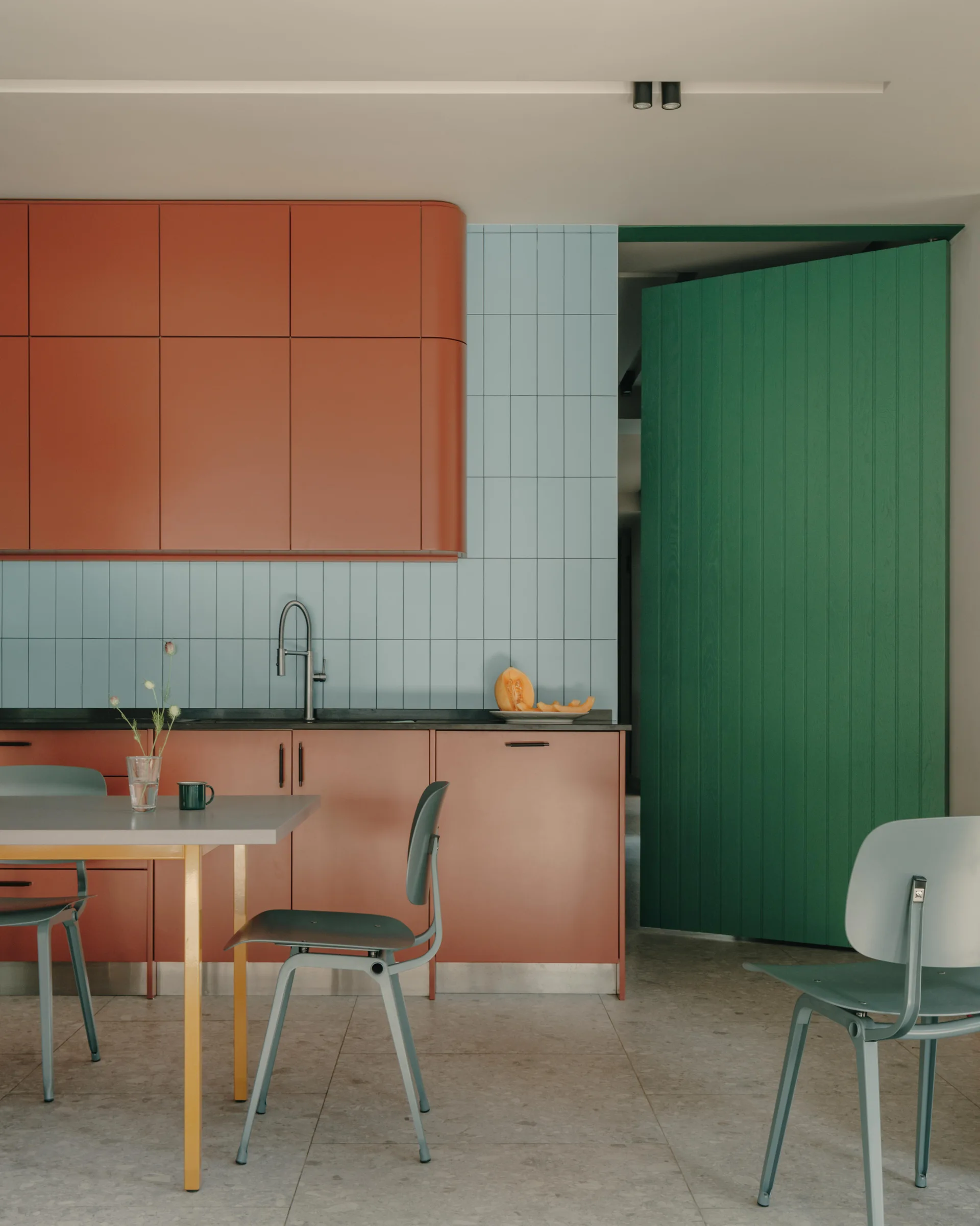 A kitchen dining area features rust-colored matte cabinets, a light blue vertical tile backsplash, grey chairs, and a bright green door.