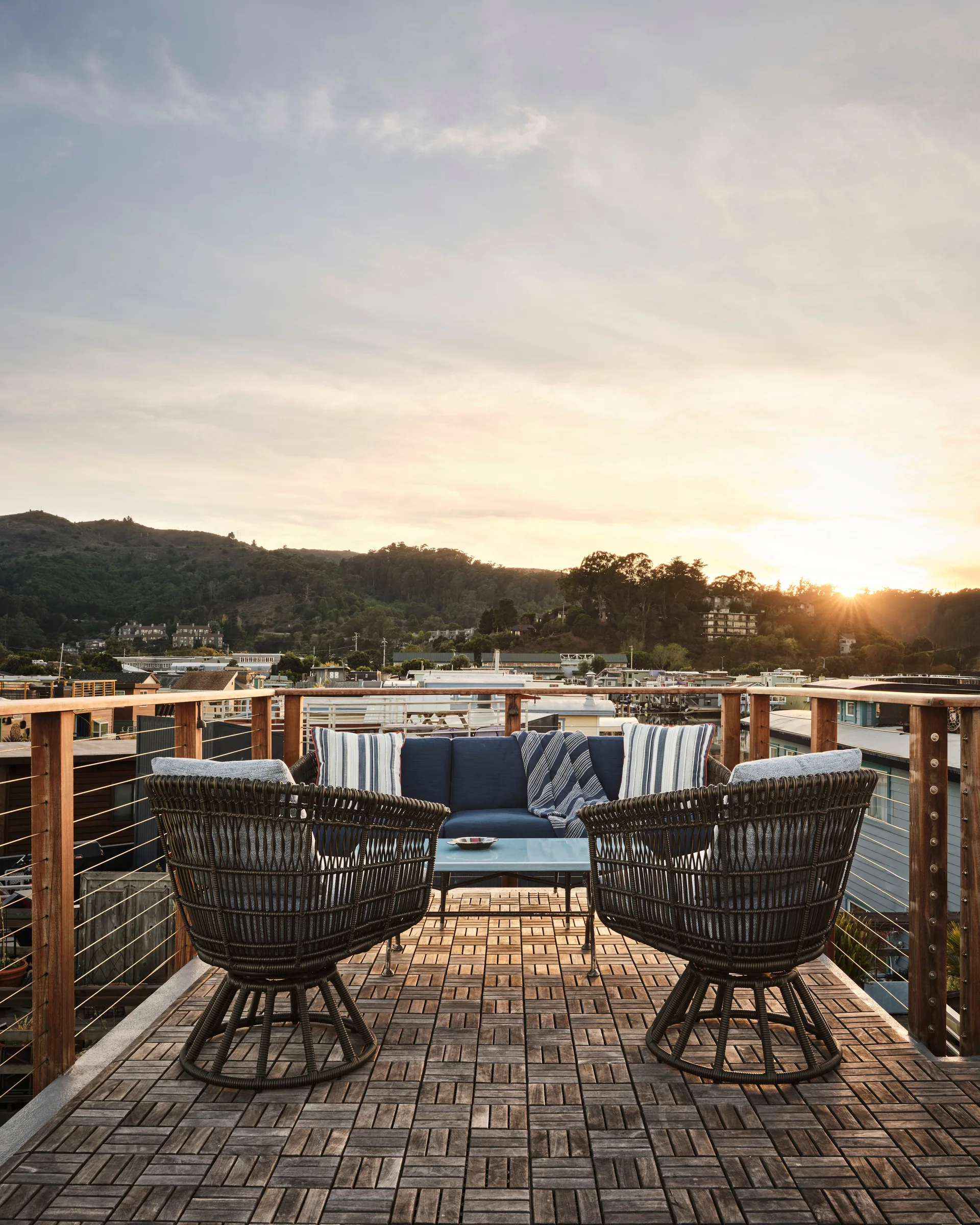 An outdoor rooftop terrace features a navy blue sofa and two woven wicker armchairs on patterned wood deck tiles overlooking a bay at sunset.