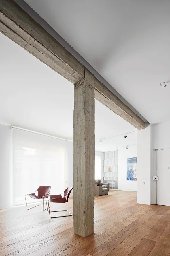 A minimalist open-plan living room featuring an exposed concrete column and beam, light wood floors, and two brown leather armchairs.