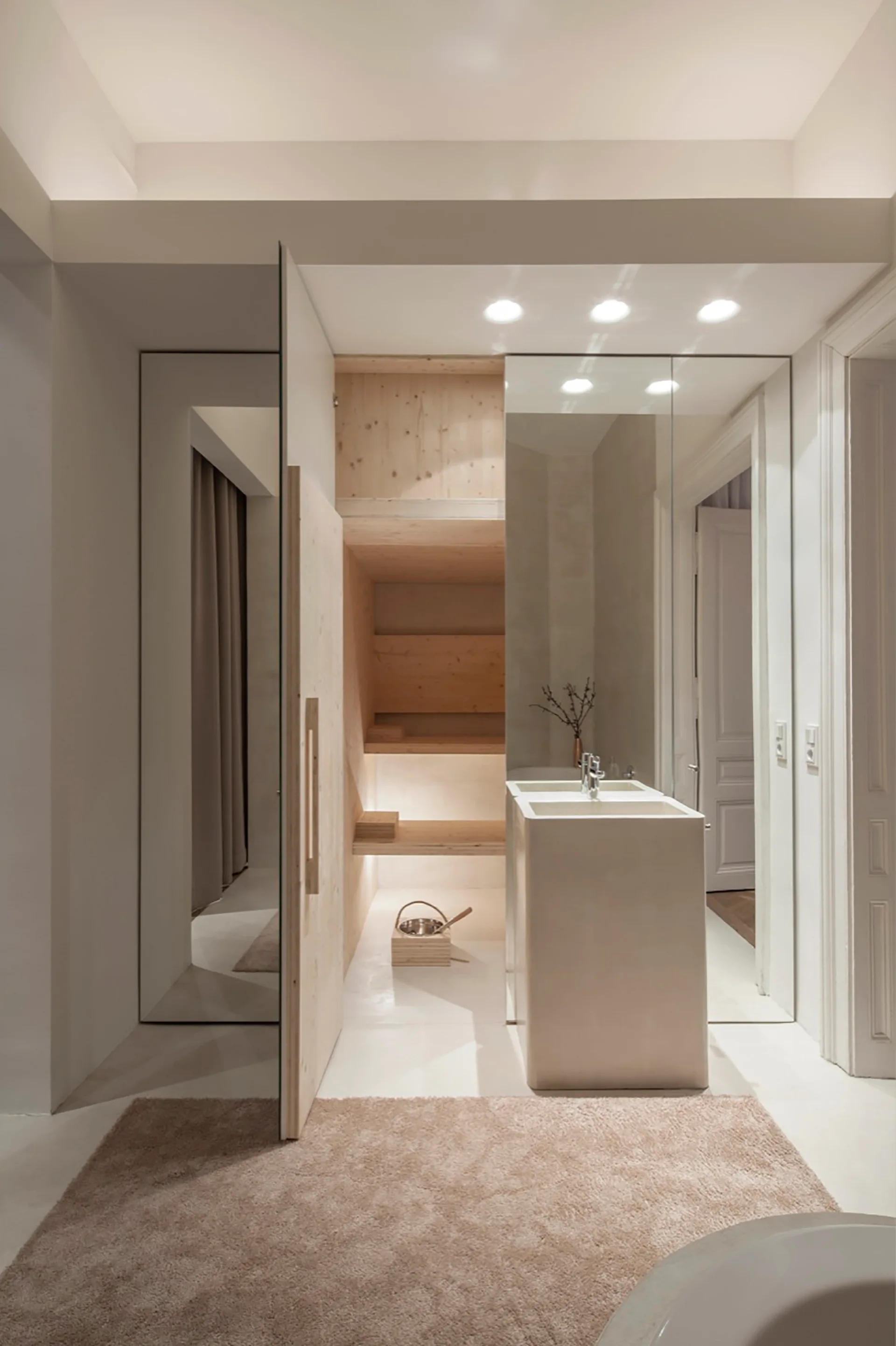 A minimalist bathroom features a freestanding monolithic pedestal sink in front of a large mirror next to a recessed light wood shelving unit.
