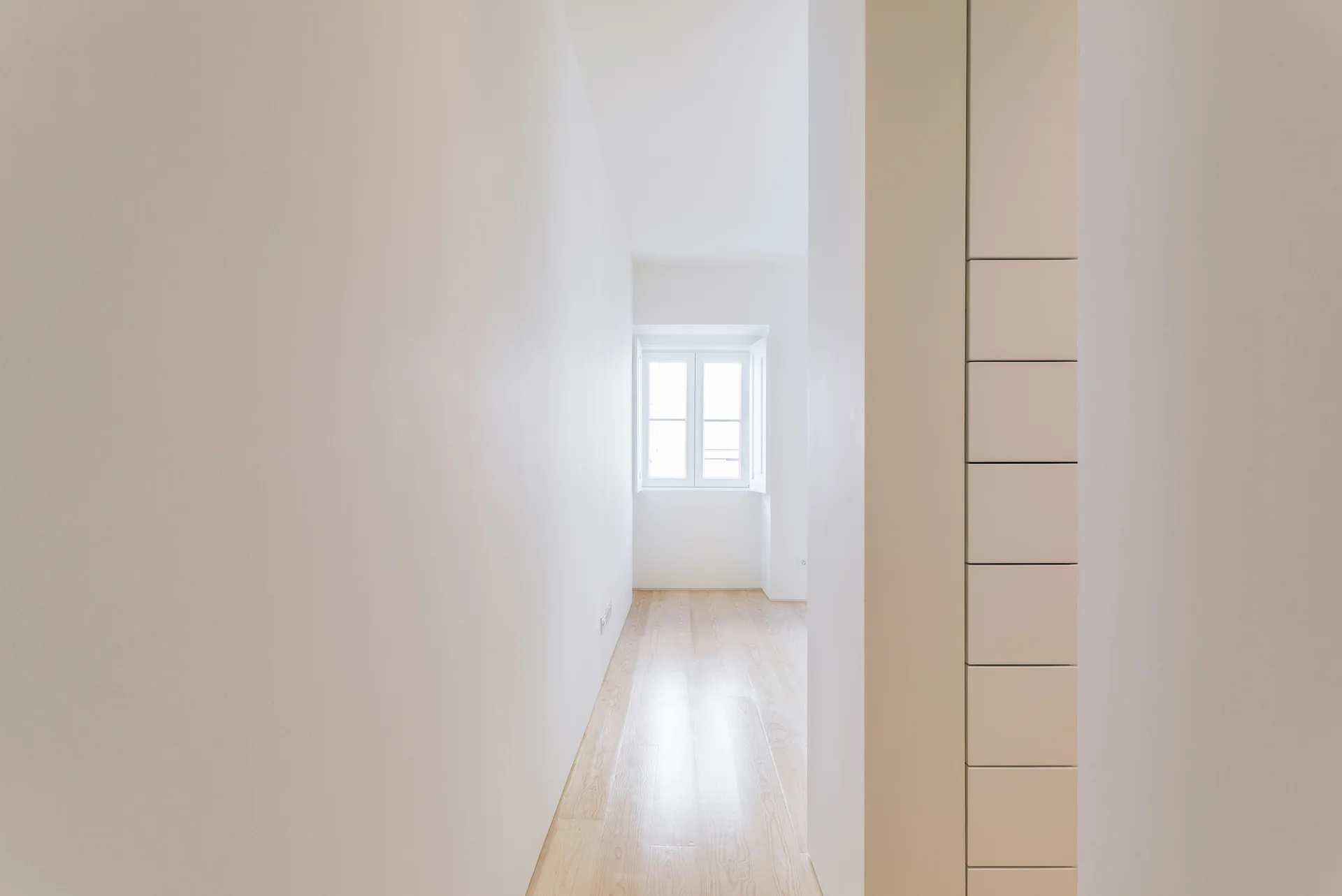 A minimalist narrow hallway featuring bright white walls, light wood plank flooring, and a single double-pane window at the far end.