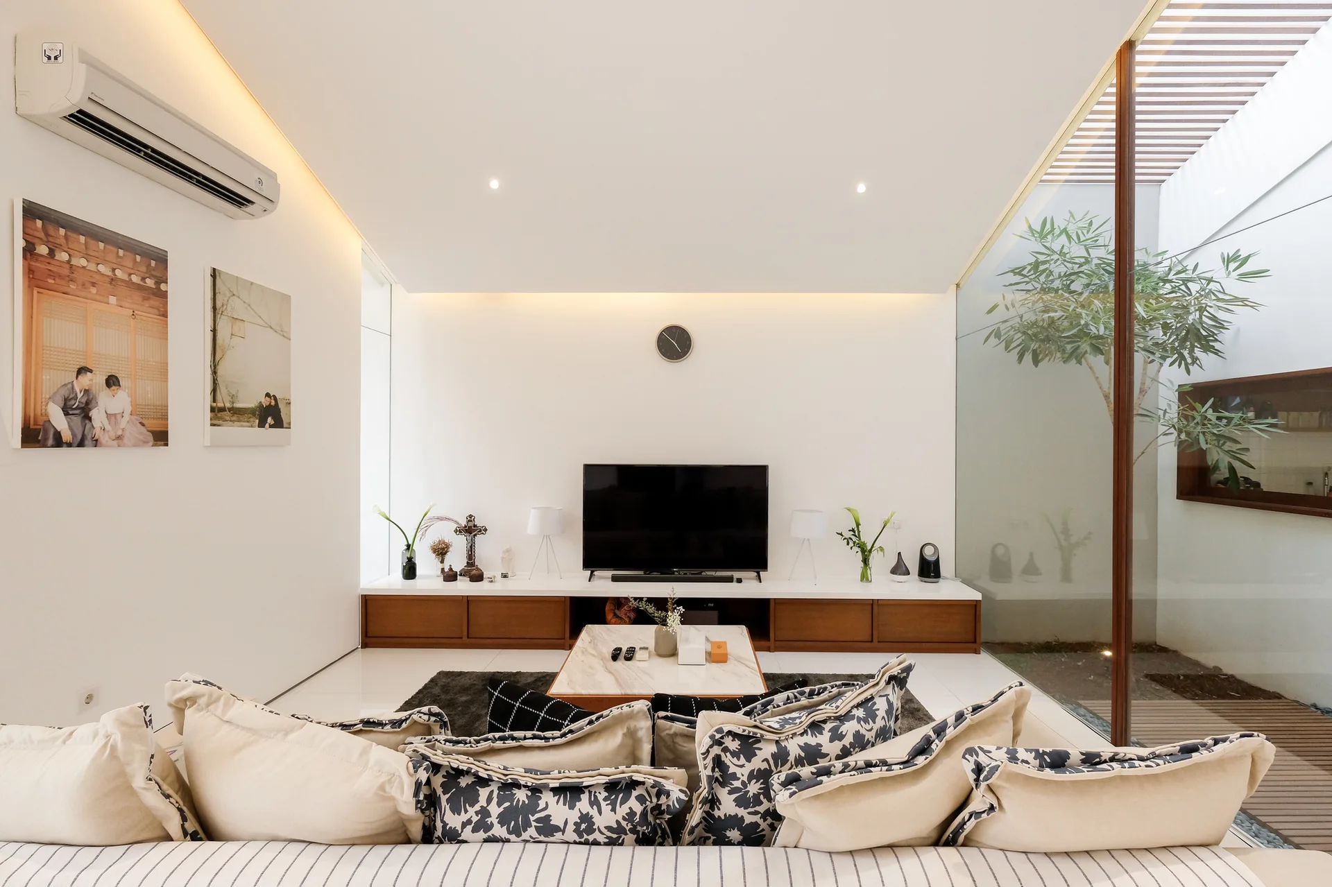 A minimalist living room with a low wood media console, a patterned sofa, and a glass wall looking out to an indoor courtyard with a small tree.