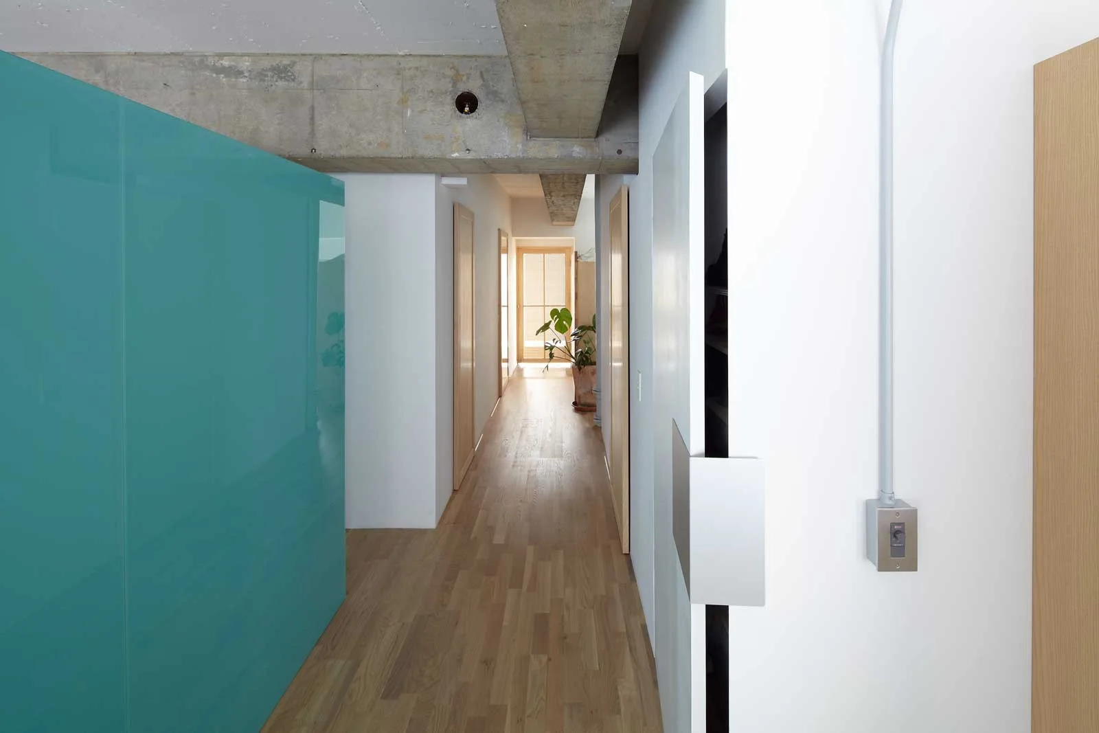 A long residential hallway features a glossy teal storage cabinet, light wood floors, and exposed raw concrete ceiling beams.