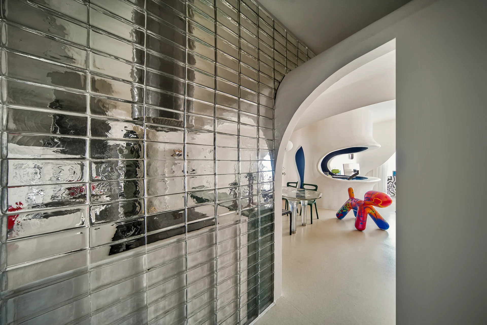 A view through a curved white archway reveals a modern dining area with a metallic glass brick wall, glass table, and colorful sculpture.